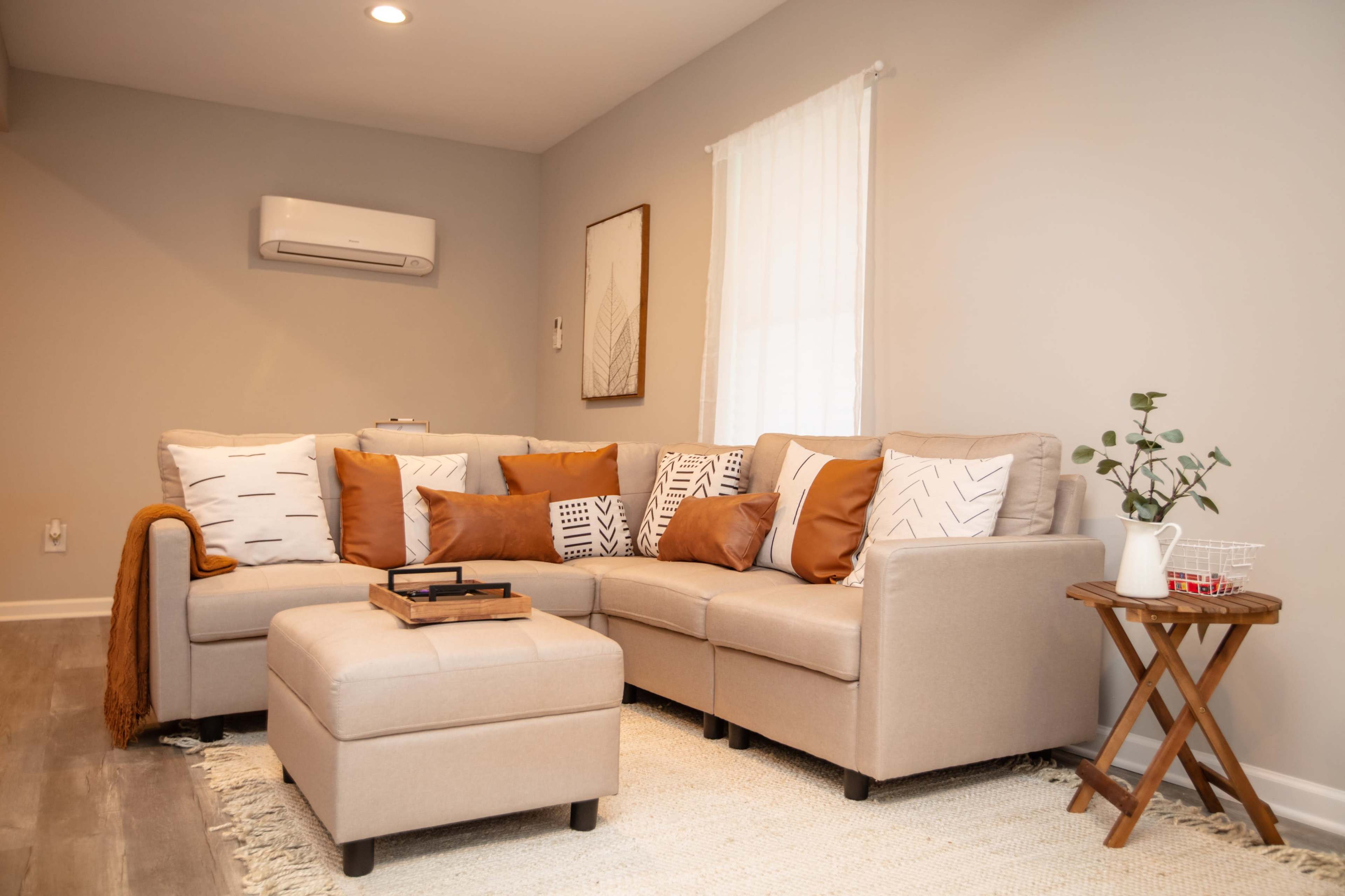 A light gray living room features a beige sectional sofa adorned with brown and patterned cushions, a small wooden side table, and a white rug.