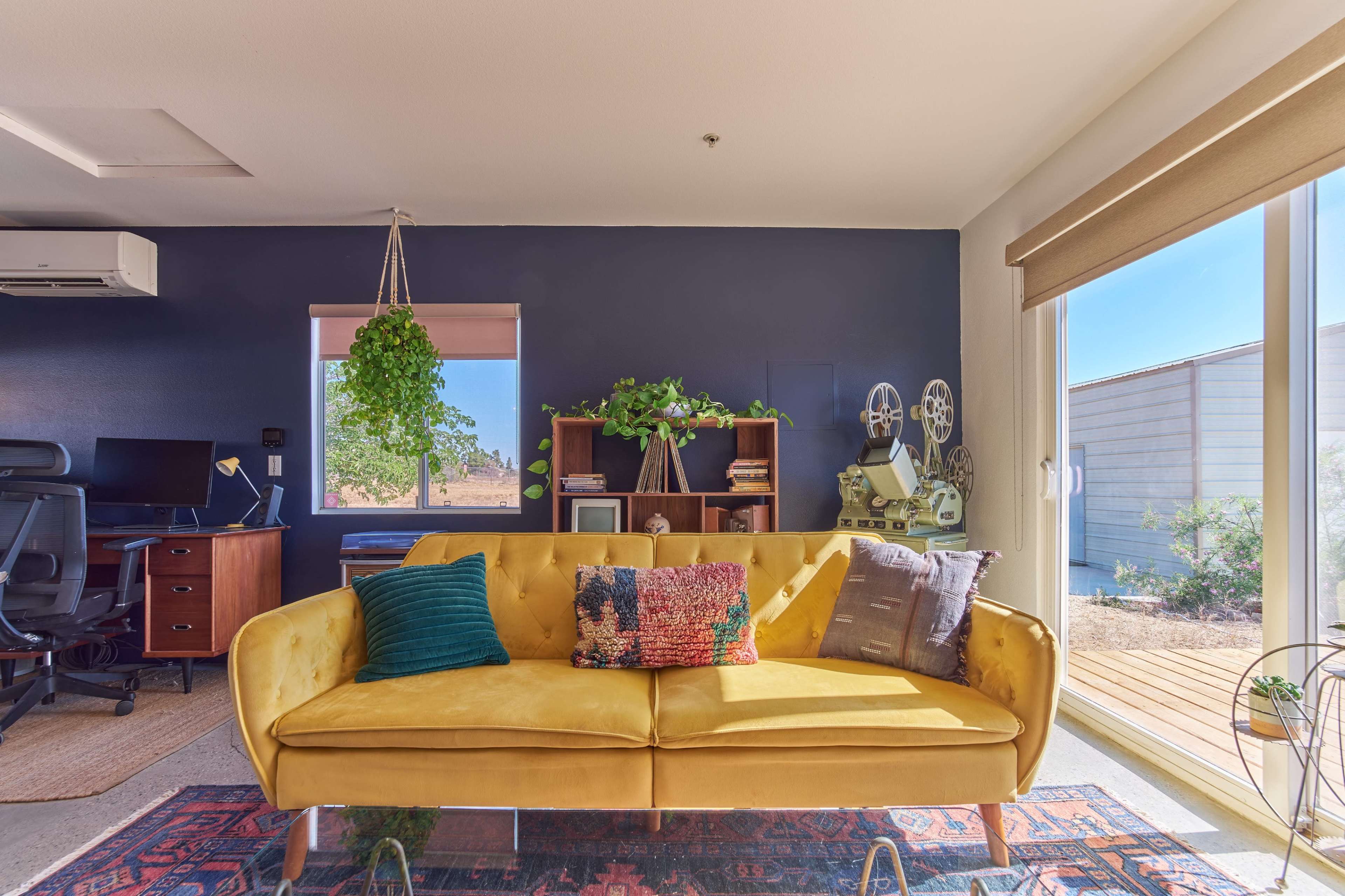 A bright yellow sofa sits in a modern workspace with a dark blue accent wall and a view of a wooden deck through large sliding glass doors.