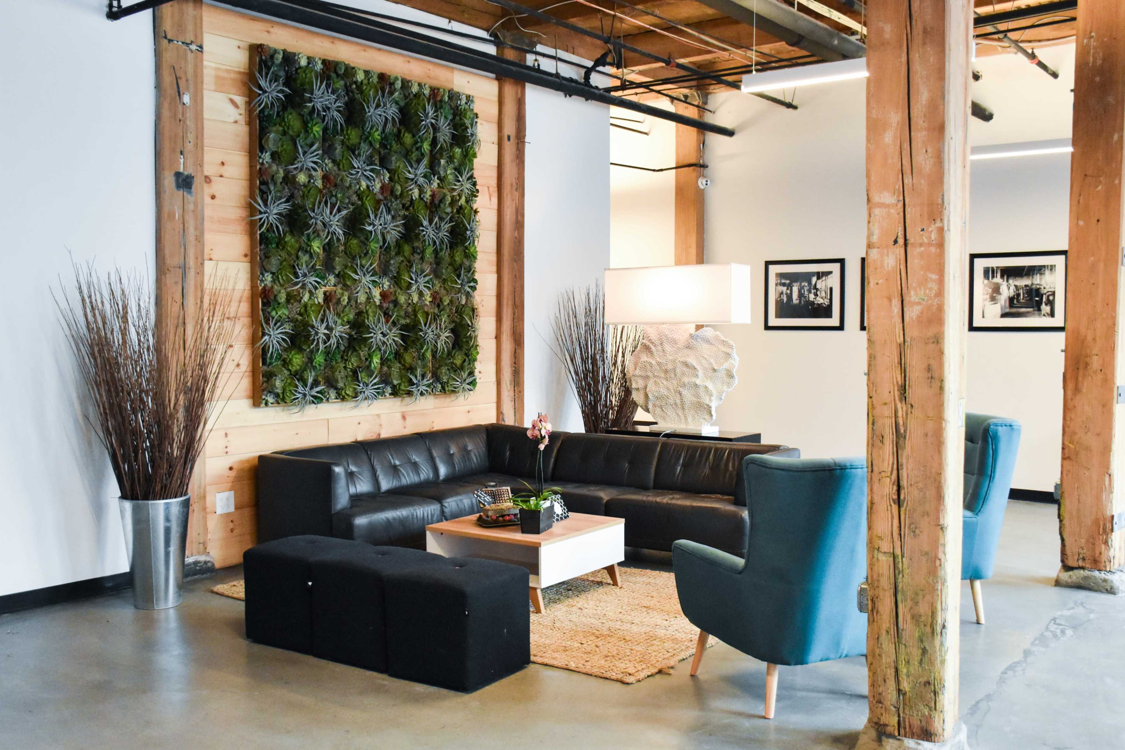 The scene features a modern lounge area with a black leather sofa, teal armchairs, a wooden coffee table, and a vertical wall of greenery, all set against a backdrop of exposed wooden beams.
