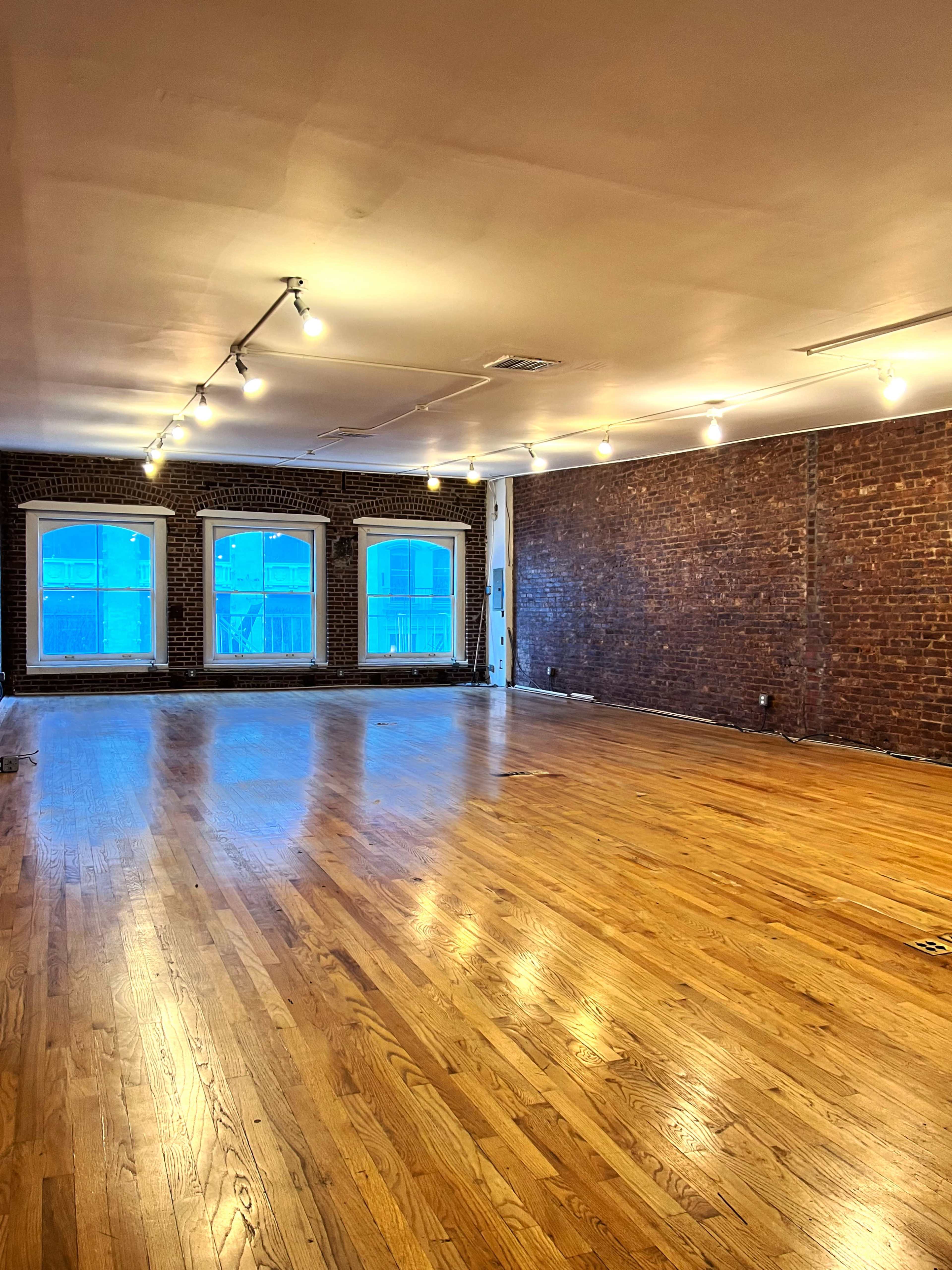A spacious, empty room with hardwood floors and exposed brick walls, featuring large windows that let in natural light.
