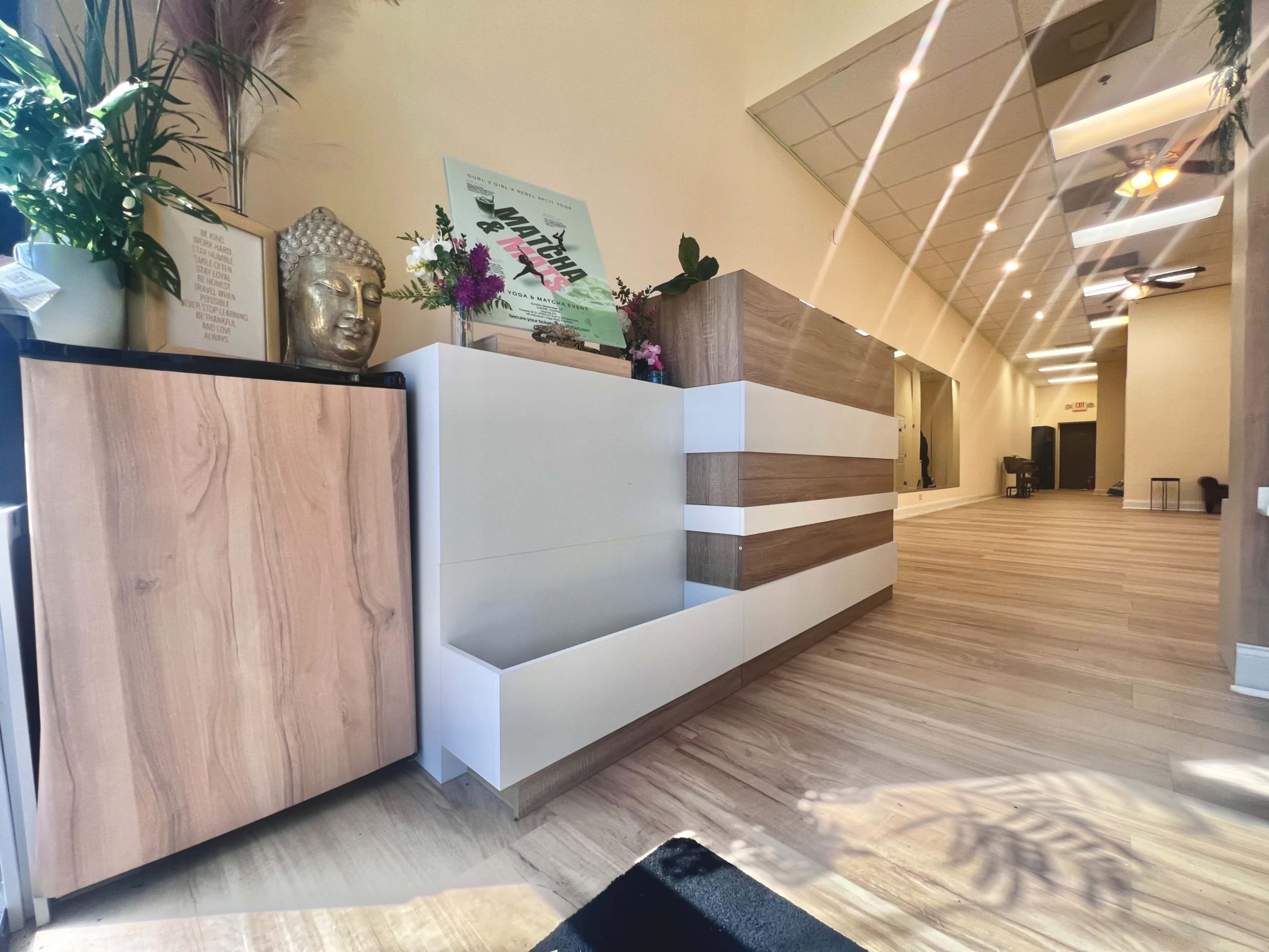 The image shows a modern interior space with light wooden flooring, a reception area featuring a mix of cabinets, decorative plants, and a Buddha statue.