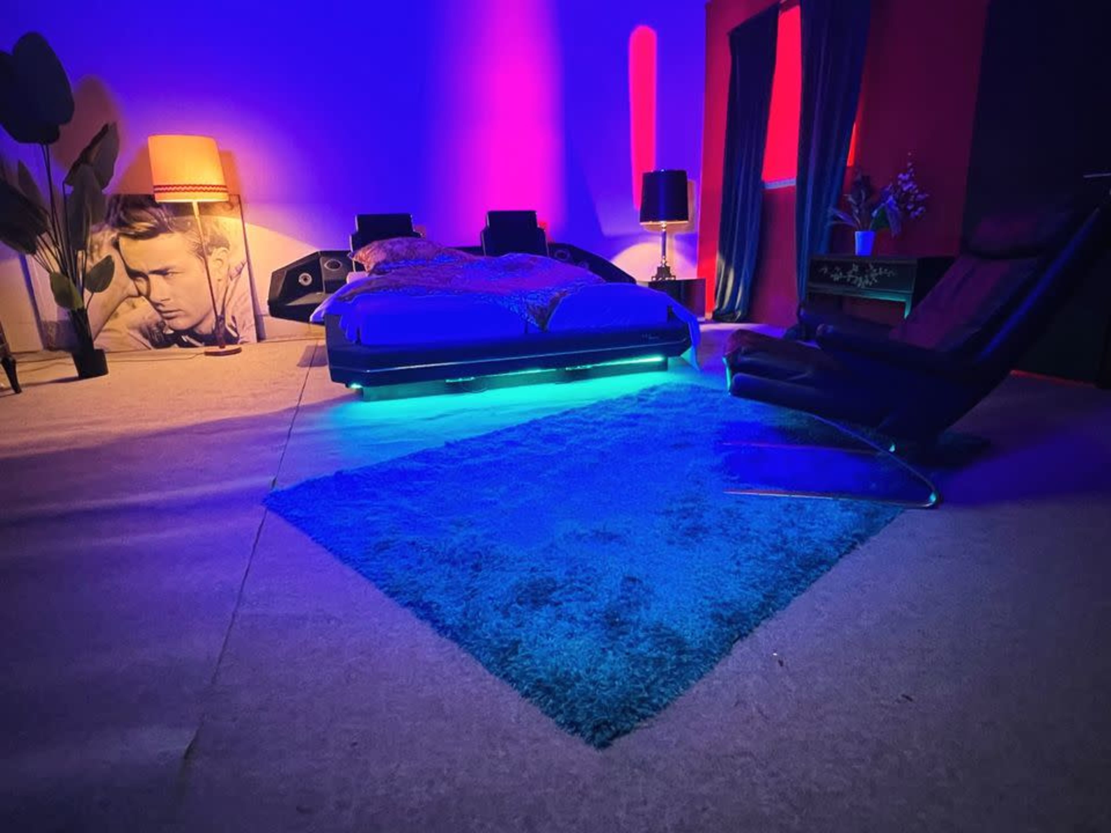 A modern bedroom with a bed illuminated by blue LED lights, a plush rug, and a black recliner chair.