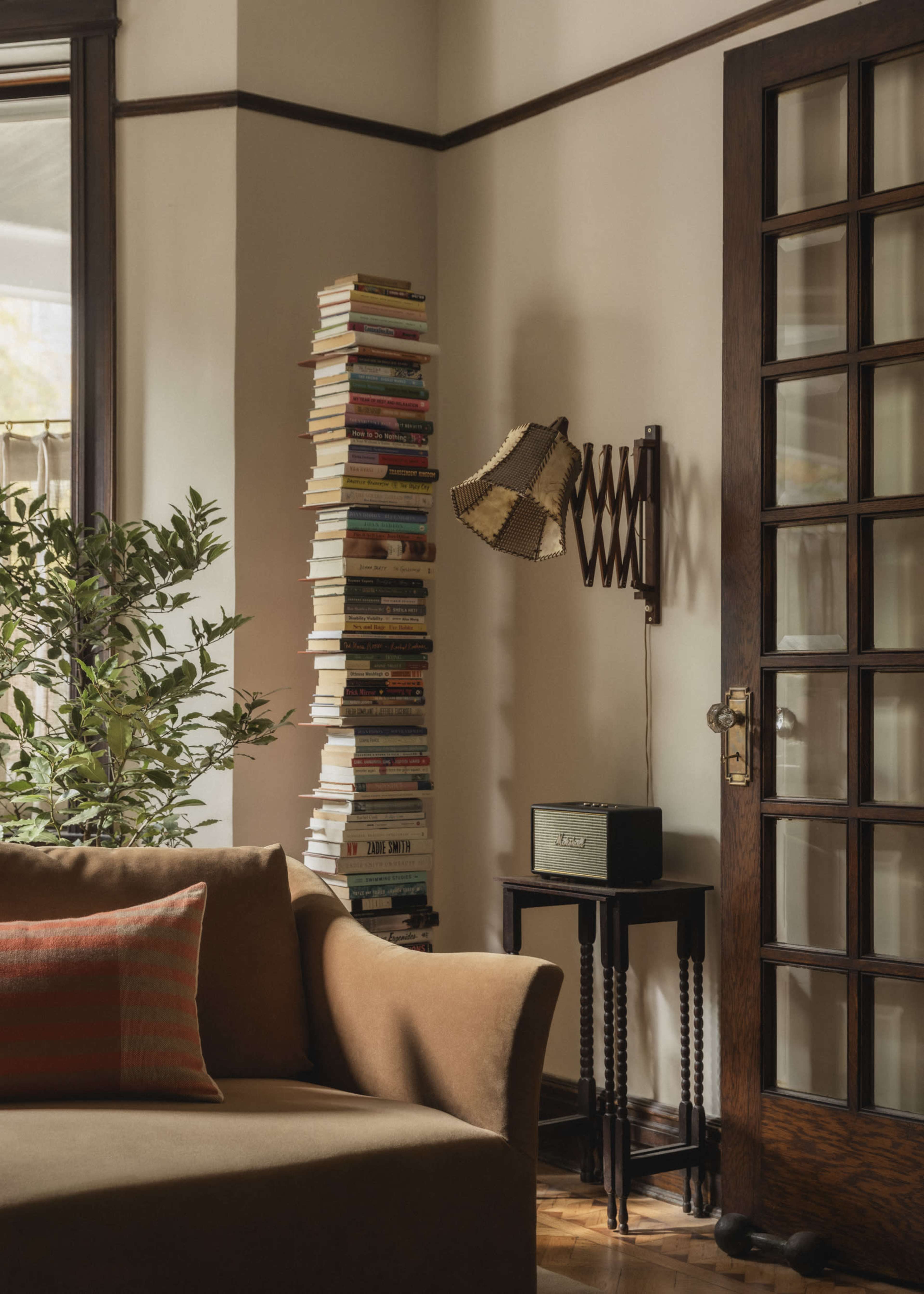 The image shows a cozy living room corner featuring a tall stack of books, a floor lamp, a small speaker on a stand, and a sofa with a decorative pillow.