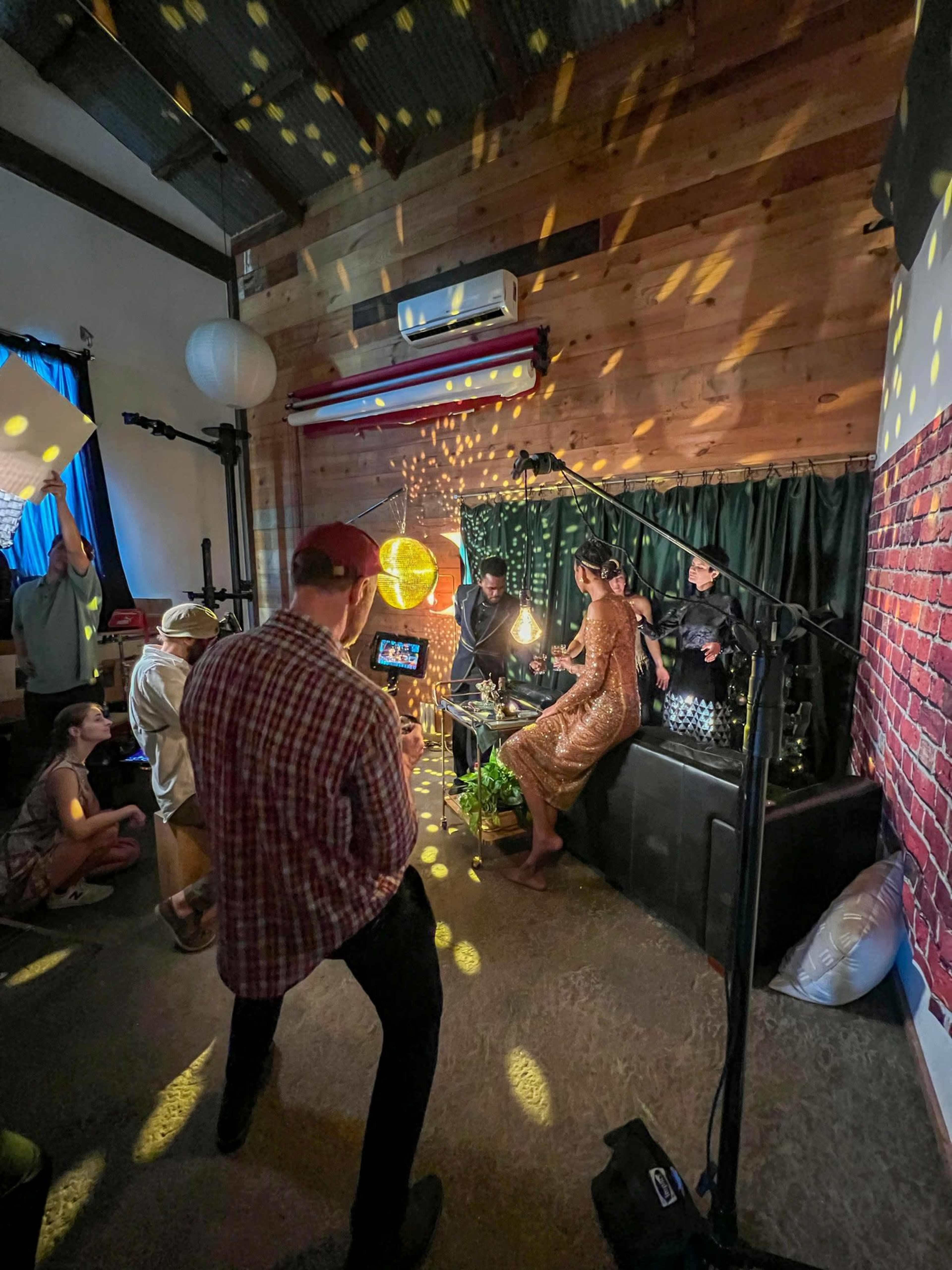 A group of people is gathered in a dimly lit room with wooden walls, engaged in a photoshoot or video production involving a model in a shiny dress and decorative lighting effects.