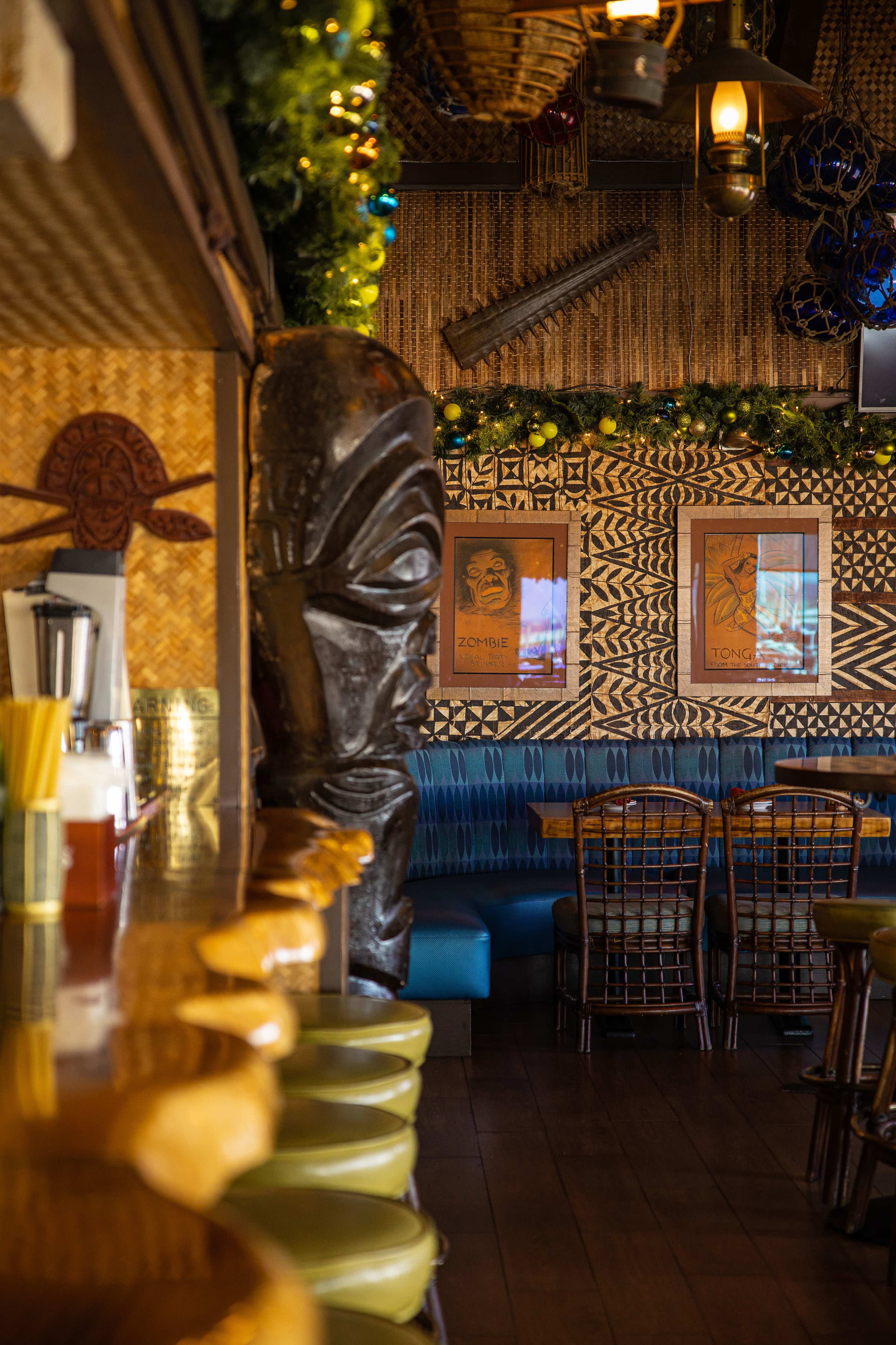 The image shows the interior of a tropical-themed restaurant featuring a tiki statue, colorful wall decorations, and a long bar with wooden stools.