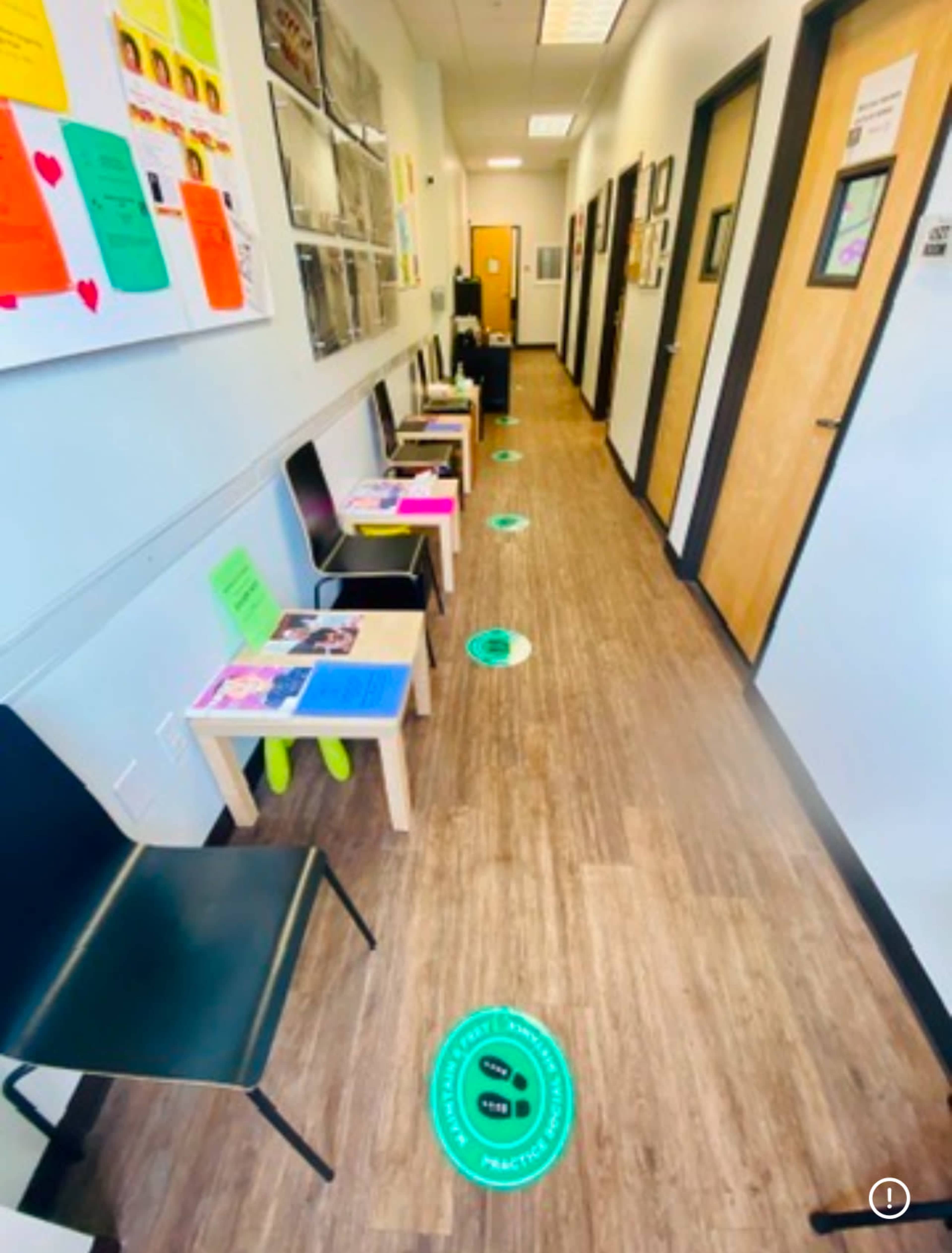 The image depicts a hallway with several chairs and small tables arranged along the sides, along with posters and signs attached to the walls.