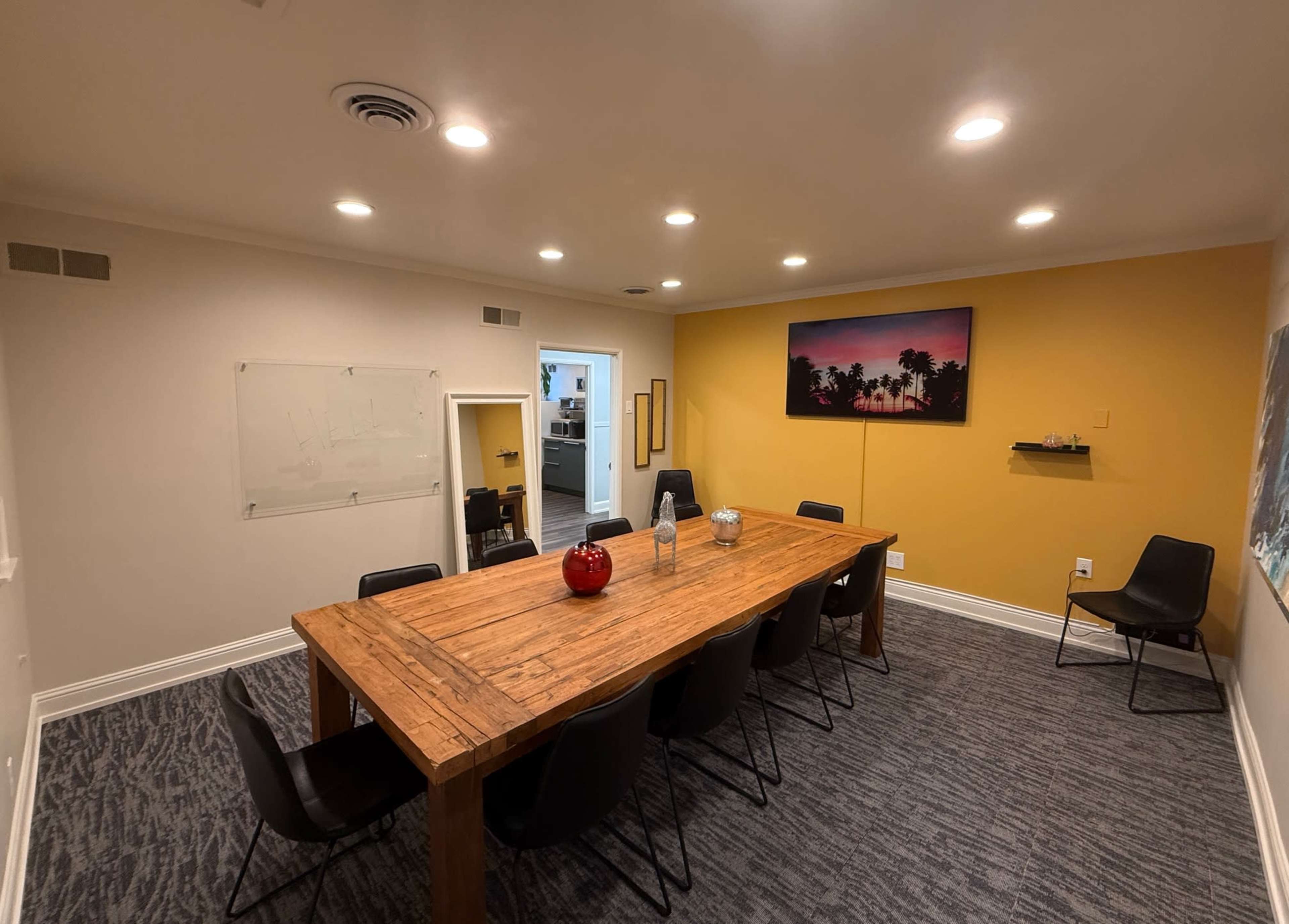 A conference room features a large wooden table surrounded by black chairs, with a yellow accent wall and a wall-mounted screen displaying a tropical scene.