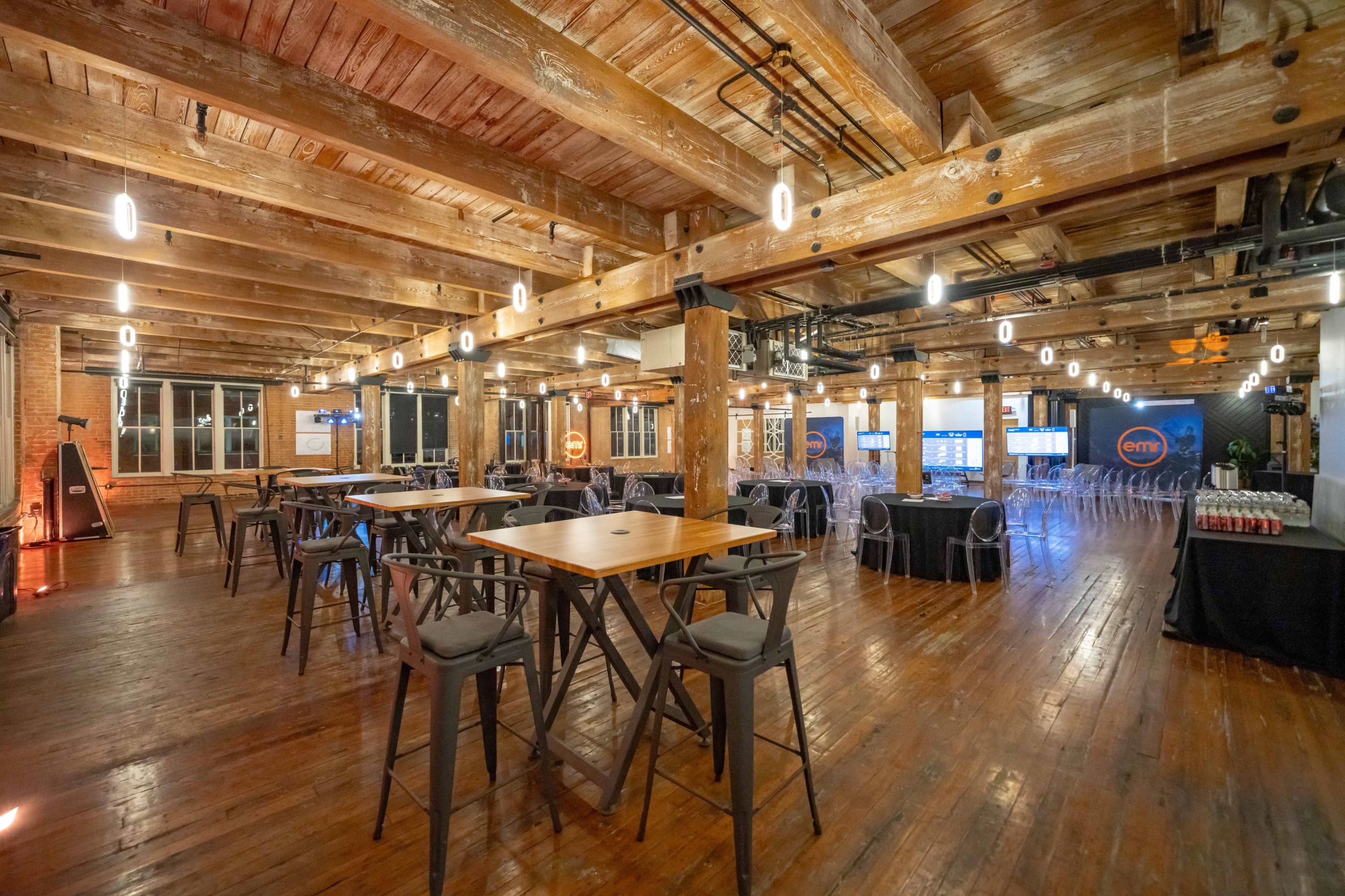 The image shows a spacious event venue with exposed wooden beams and floors, featuring tables, chairs, and screens arranged for a gathering.