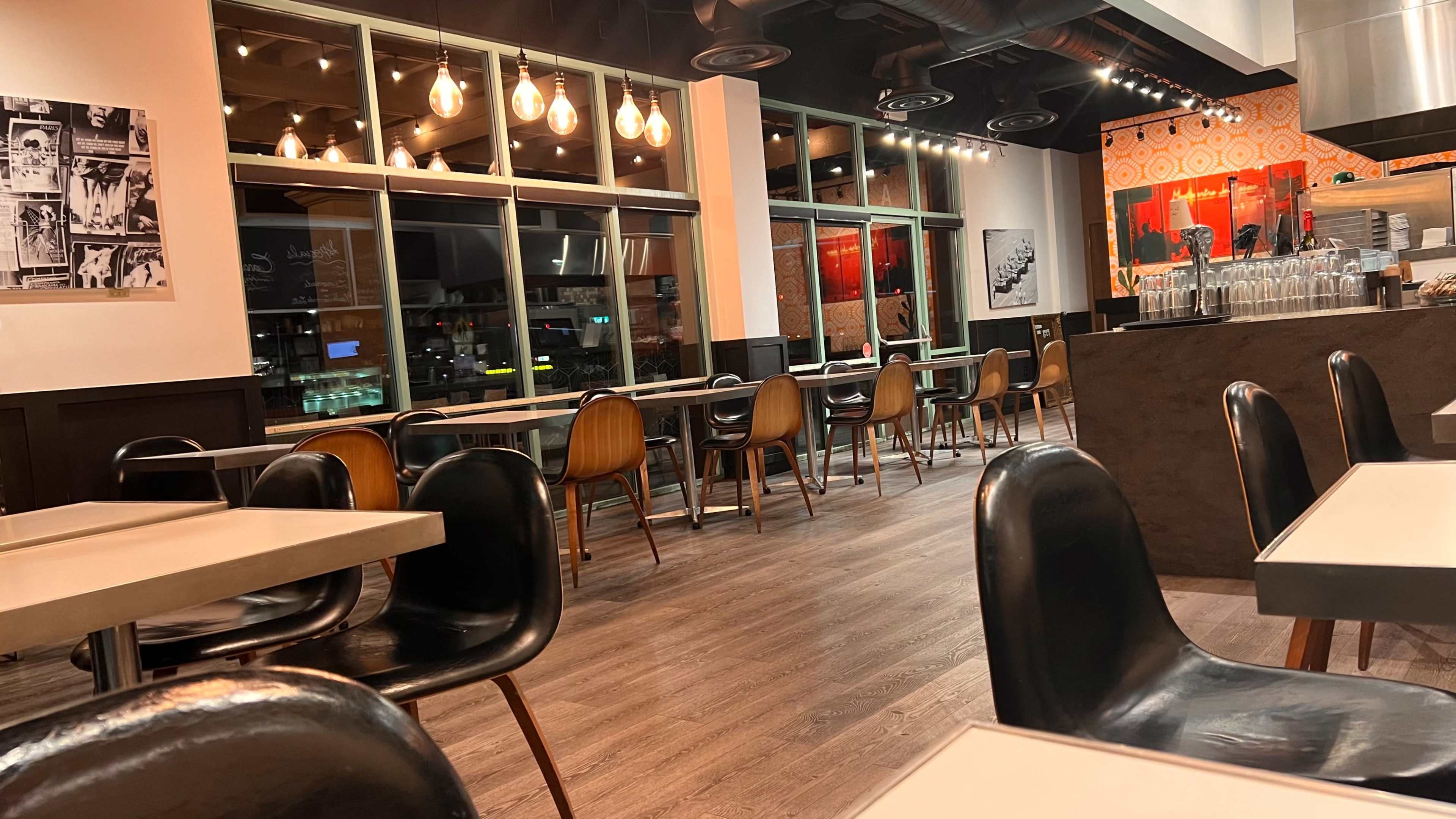 An empty restaurant interior featuring wooden flooring, a variety of chairs, and large windows with pendant lights overhead.