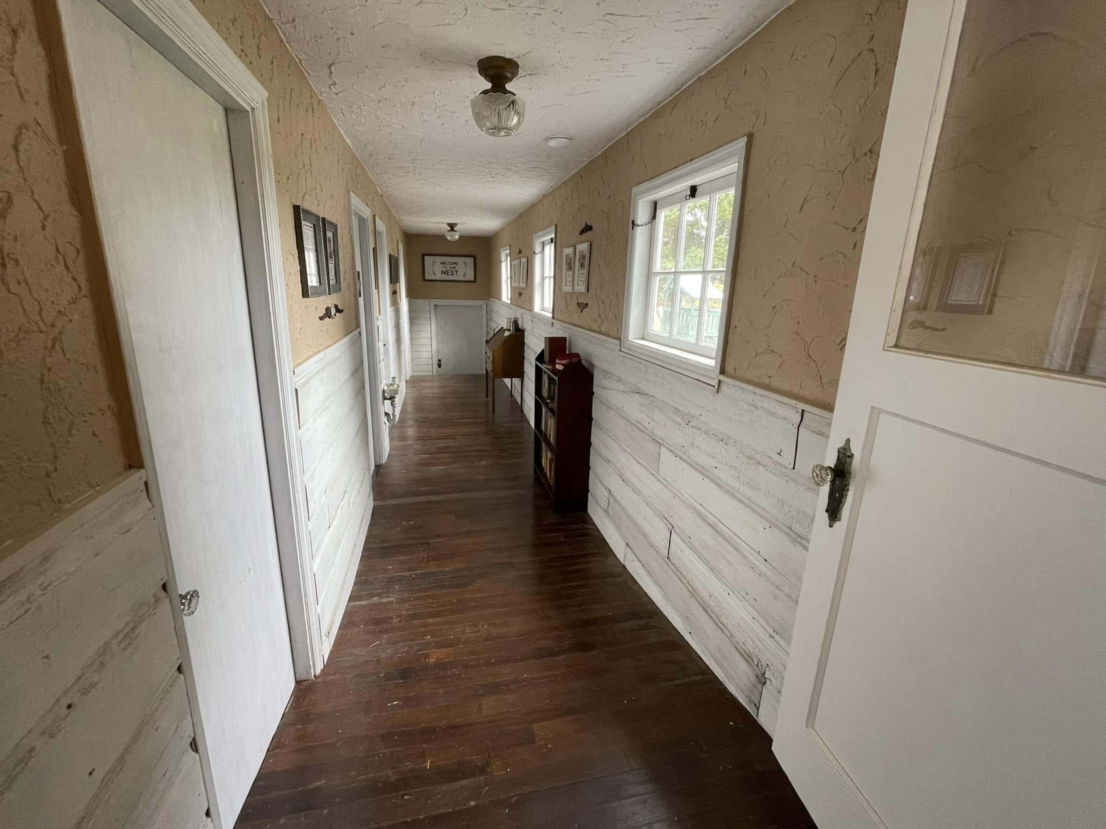 A long, narrow hallway with wooden floors and white walls, featuring several doors and a small wooden cabinet against the wall.