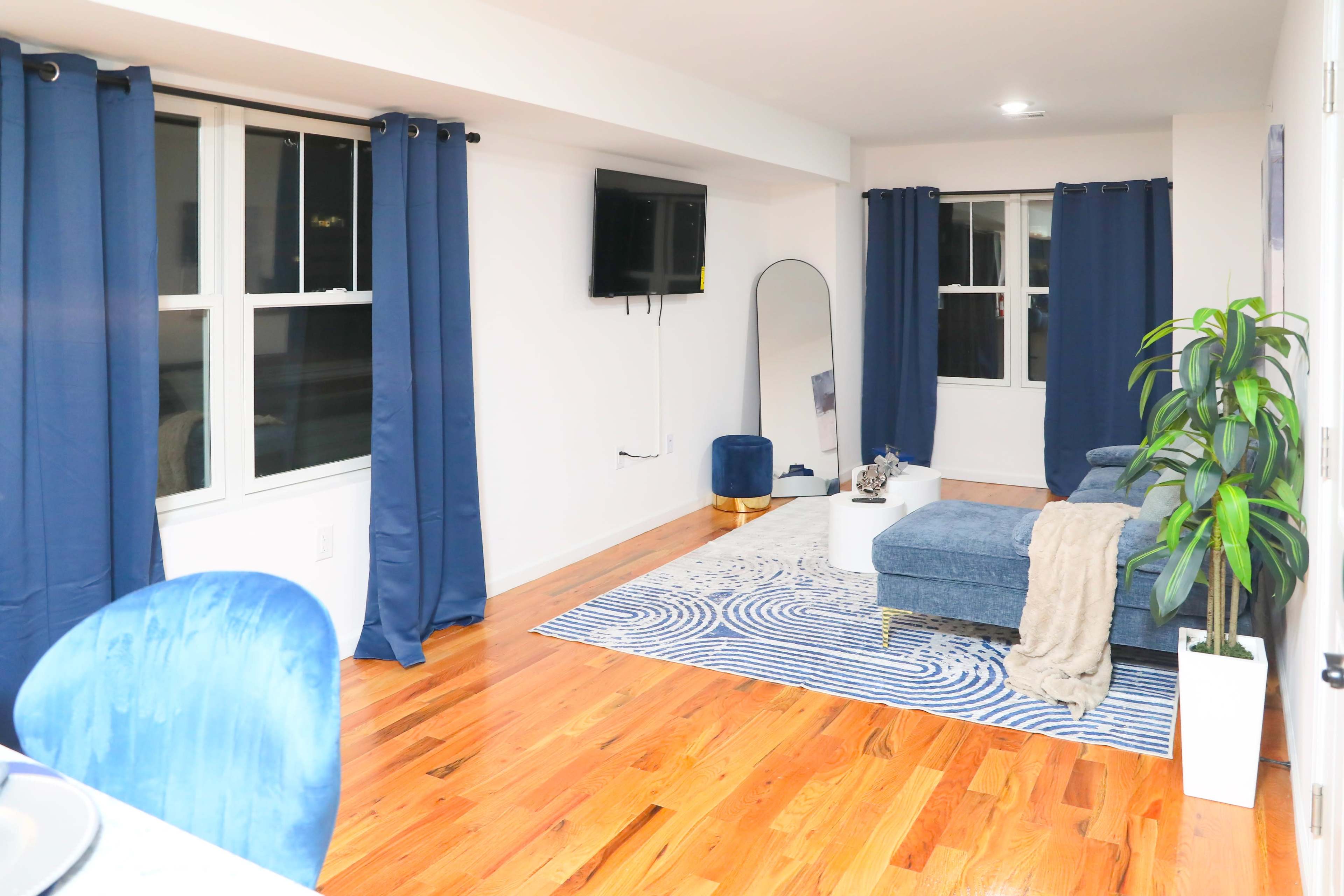 The image shows a modern living room with a blue sectional couch, a large area rug, a television mounted on the wall, and large windows with navy curtains.