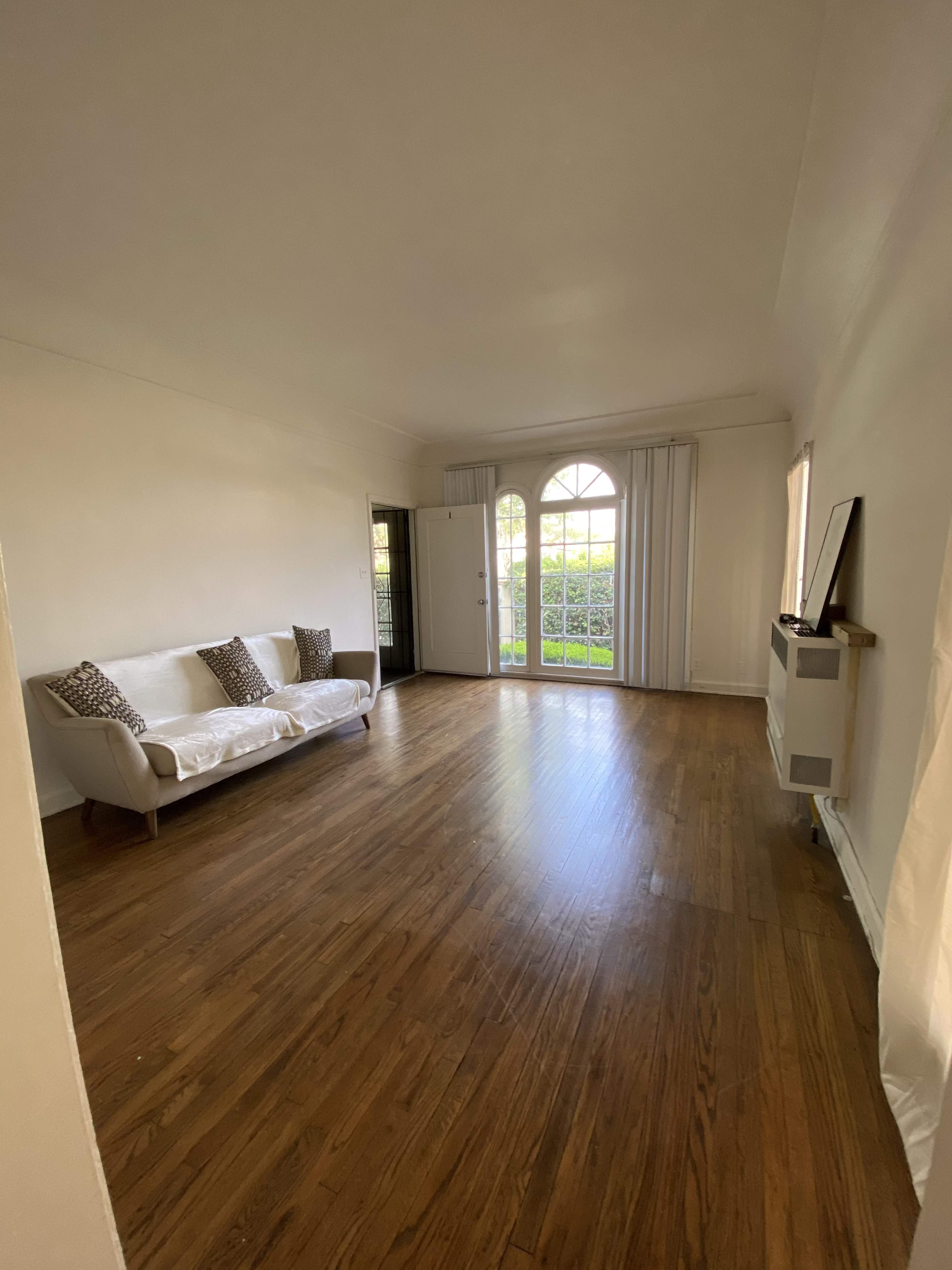 A spacious, empty living room features a white sofa and large windows with natural light.