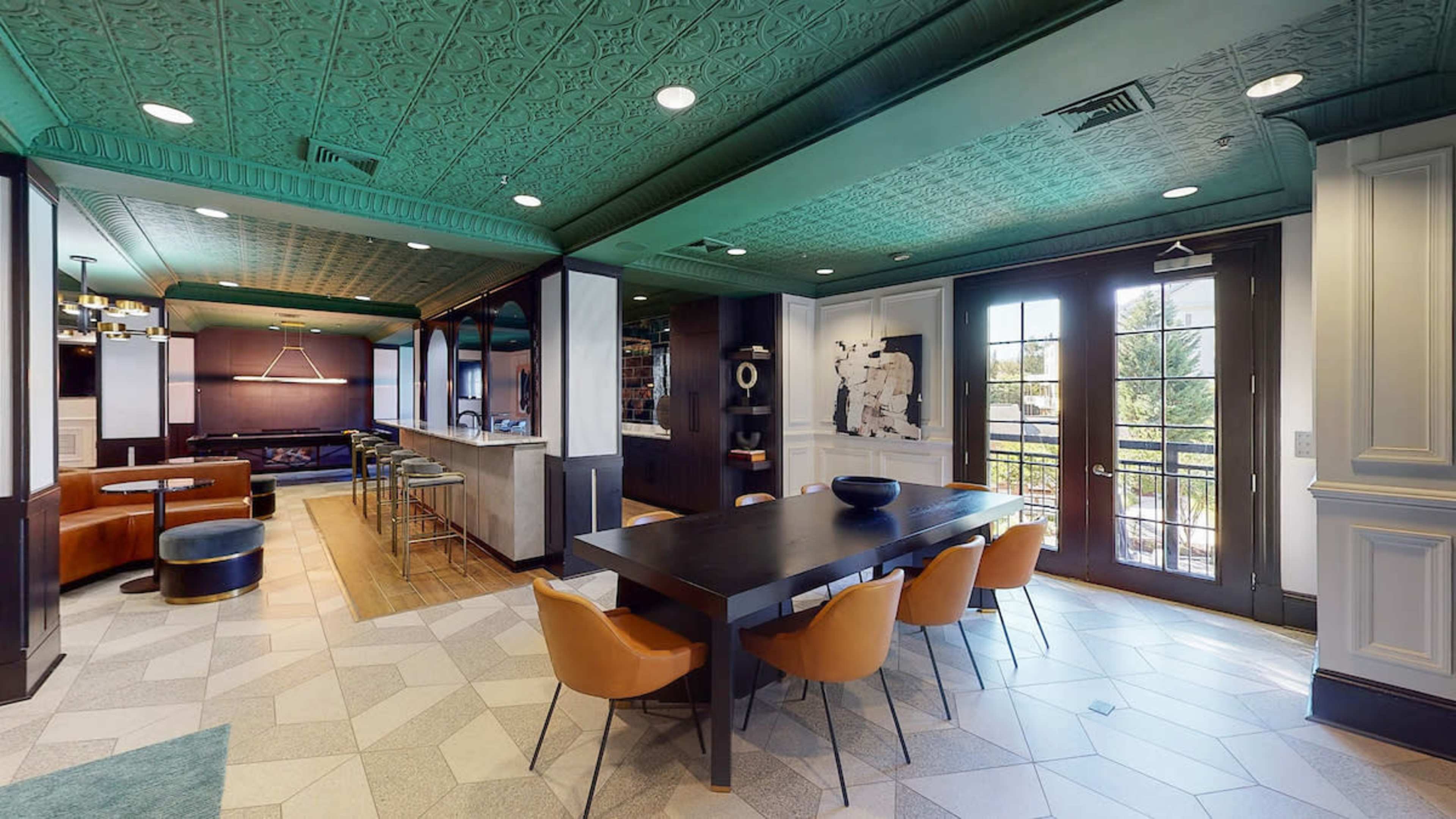 A modern lounge area featuring a dining table with orange chairs, a sleek kitchen, and large windows letting in natural light.