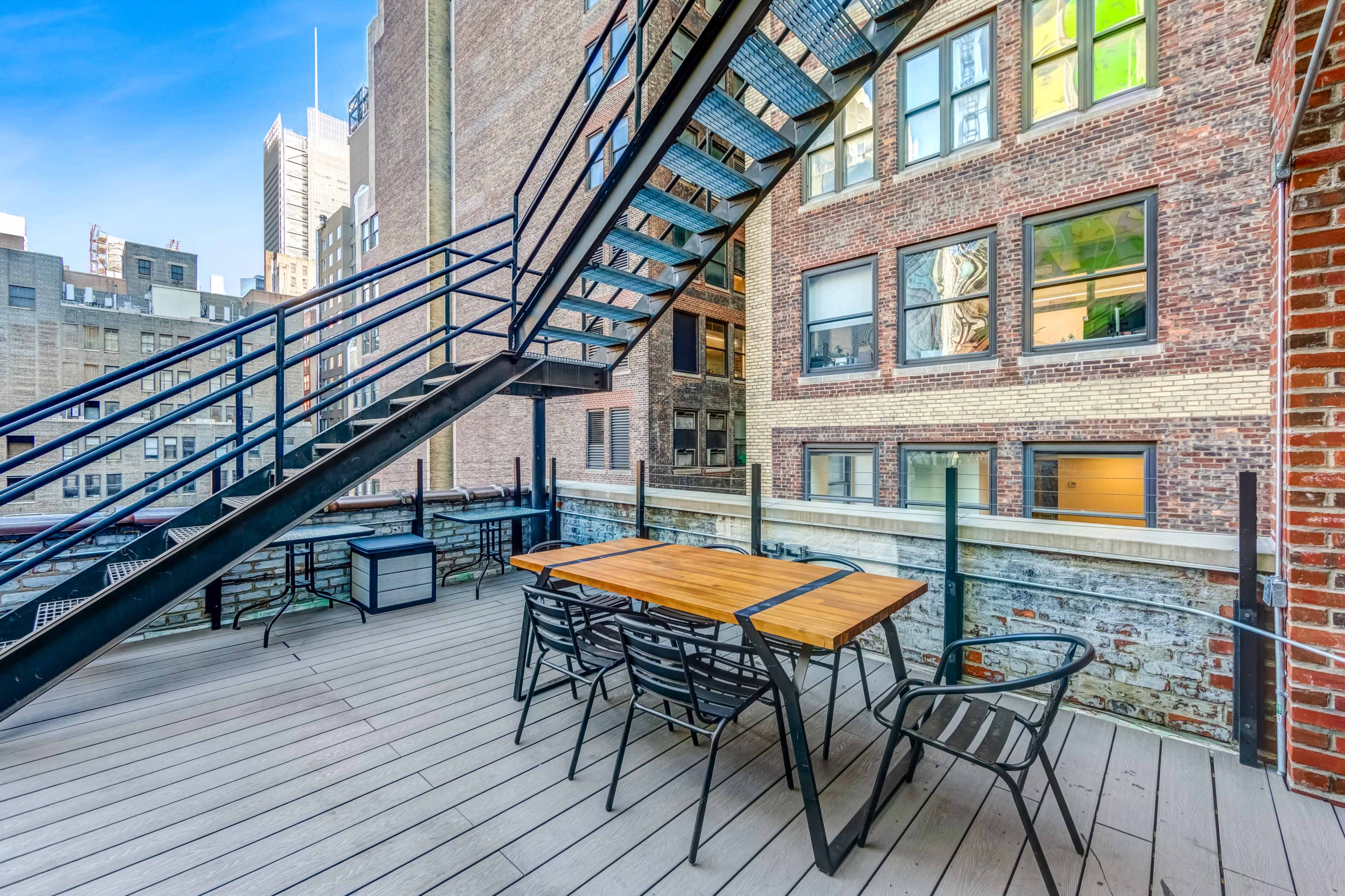 The image shows a rooftop terrace with a wooden table and chairs beneath a metal staircase, surrounded by brick buildings and large windows.