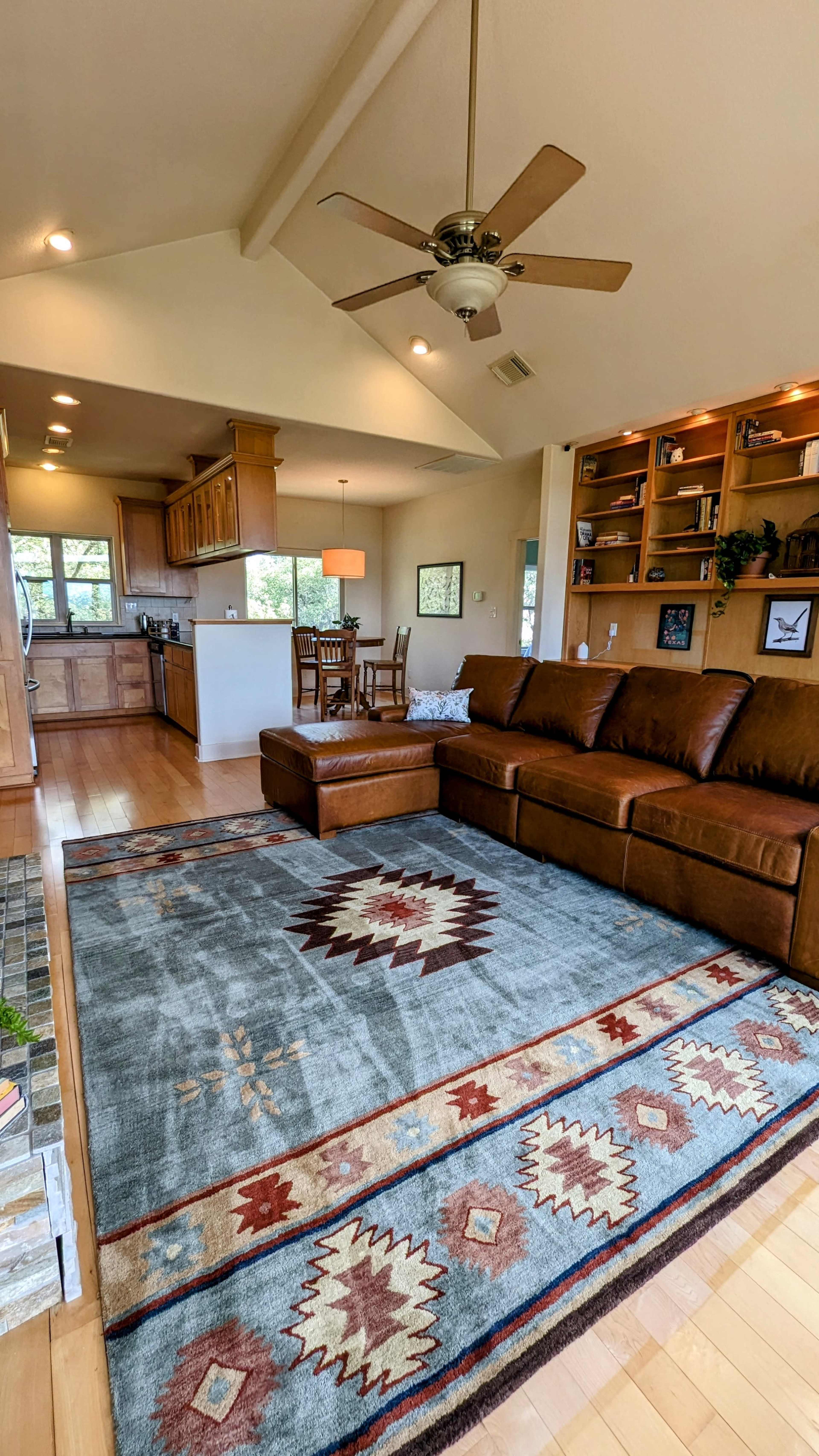 A living room with a brown sectional sofa, a patterned rug, and a visible kitchen area in the background.