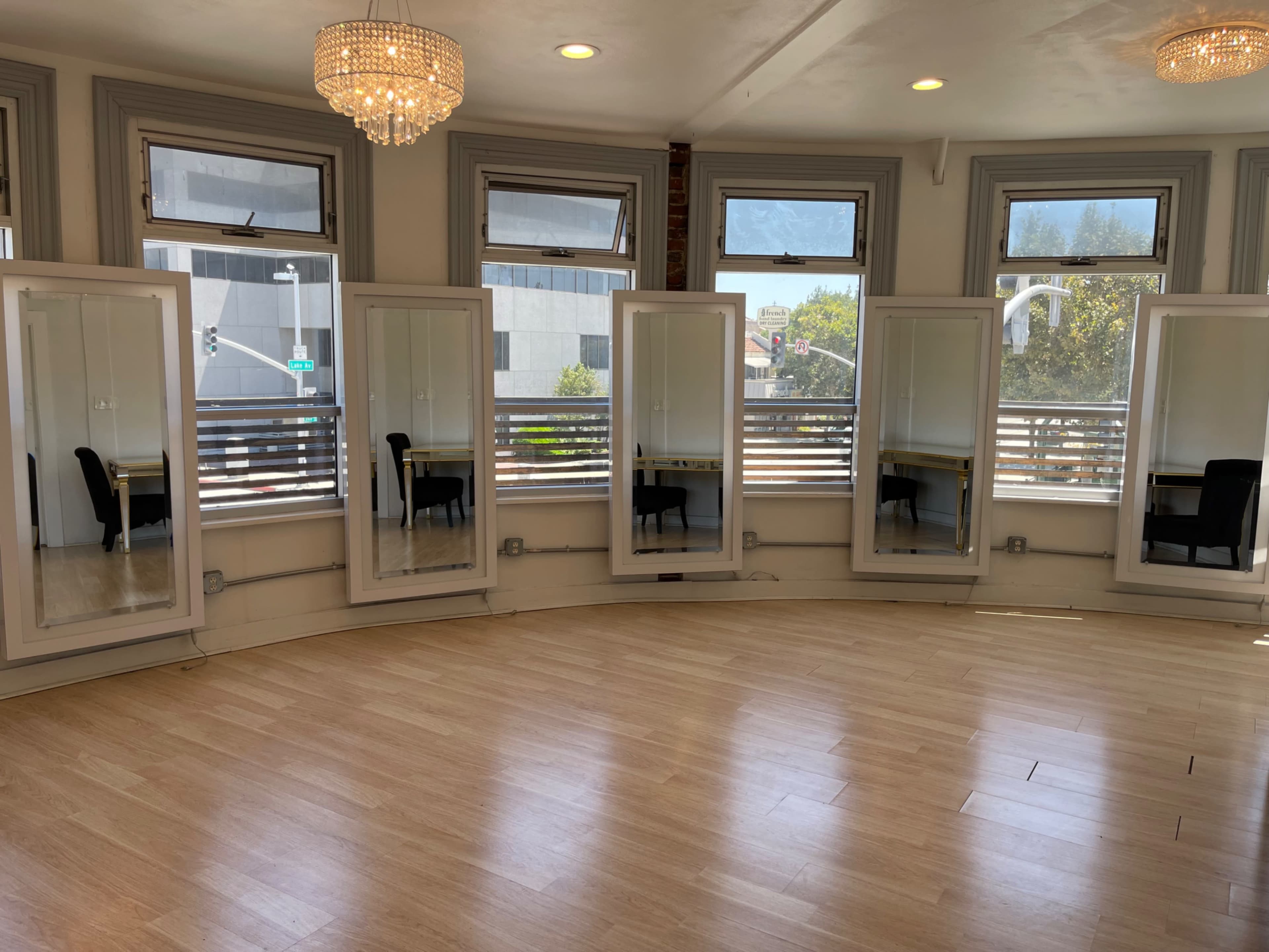 A sunlit room with wooden flooring features five tall mirrors along the curved wall and a table with a chair in the corner.