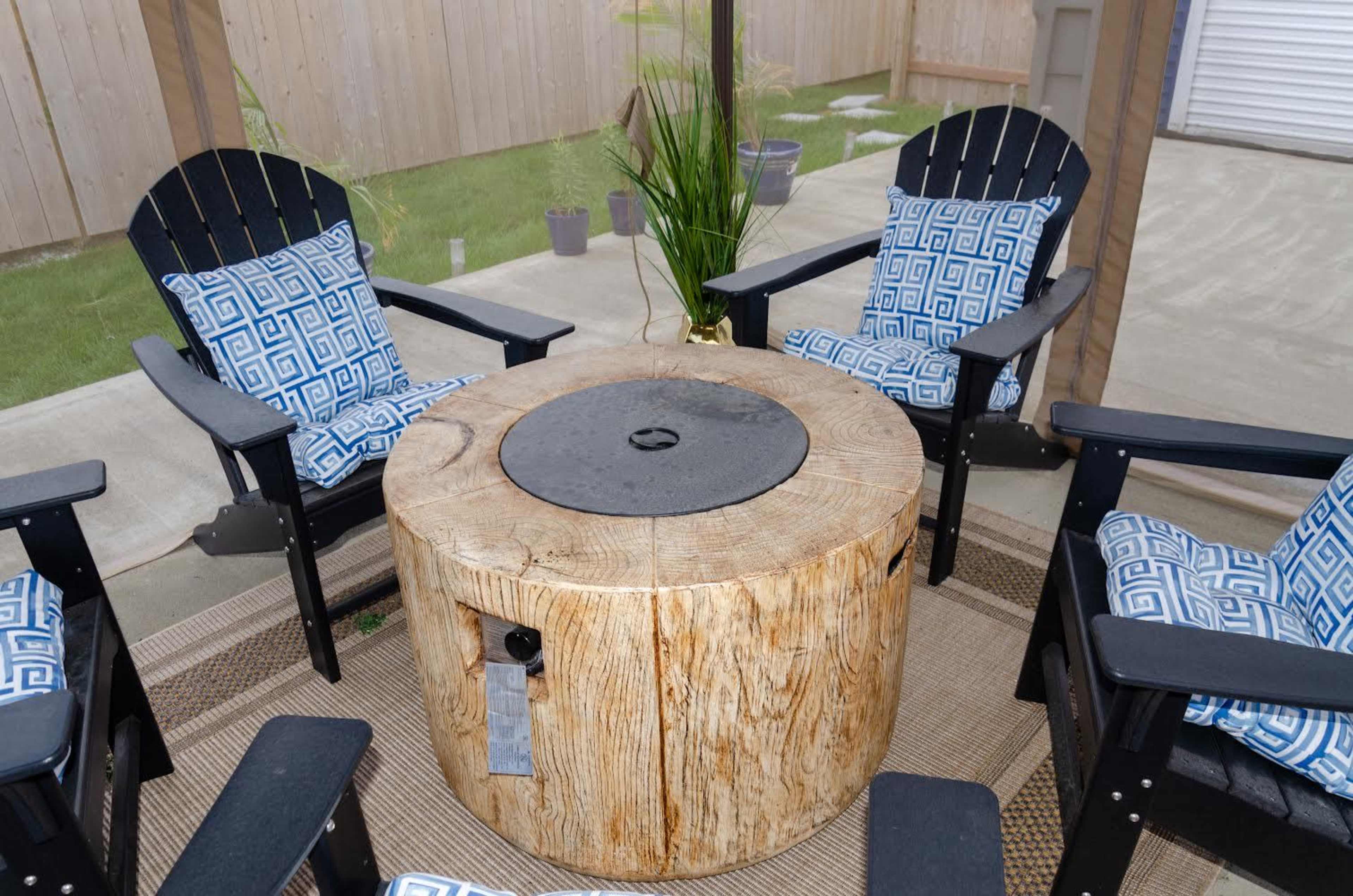 The image shows a circular wooden fire pit surrounded by four black adirondack chairs with blue-patterned cushions, all situated on a patio.