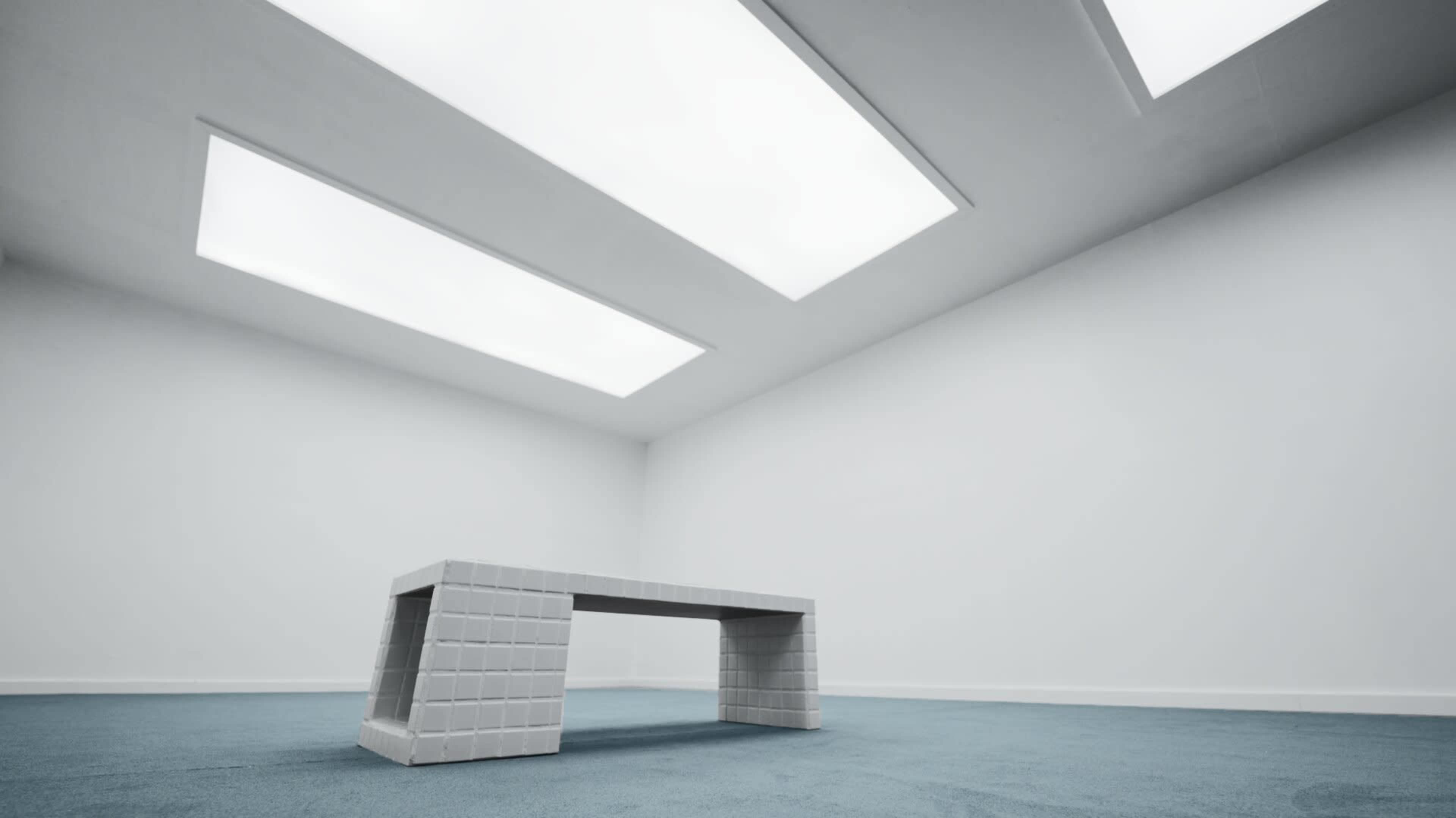 A minimalistic room features a textured white table and three overhead skylights illuminating the space.