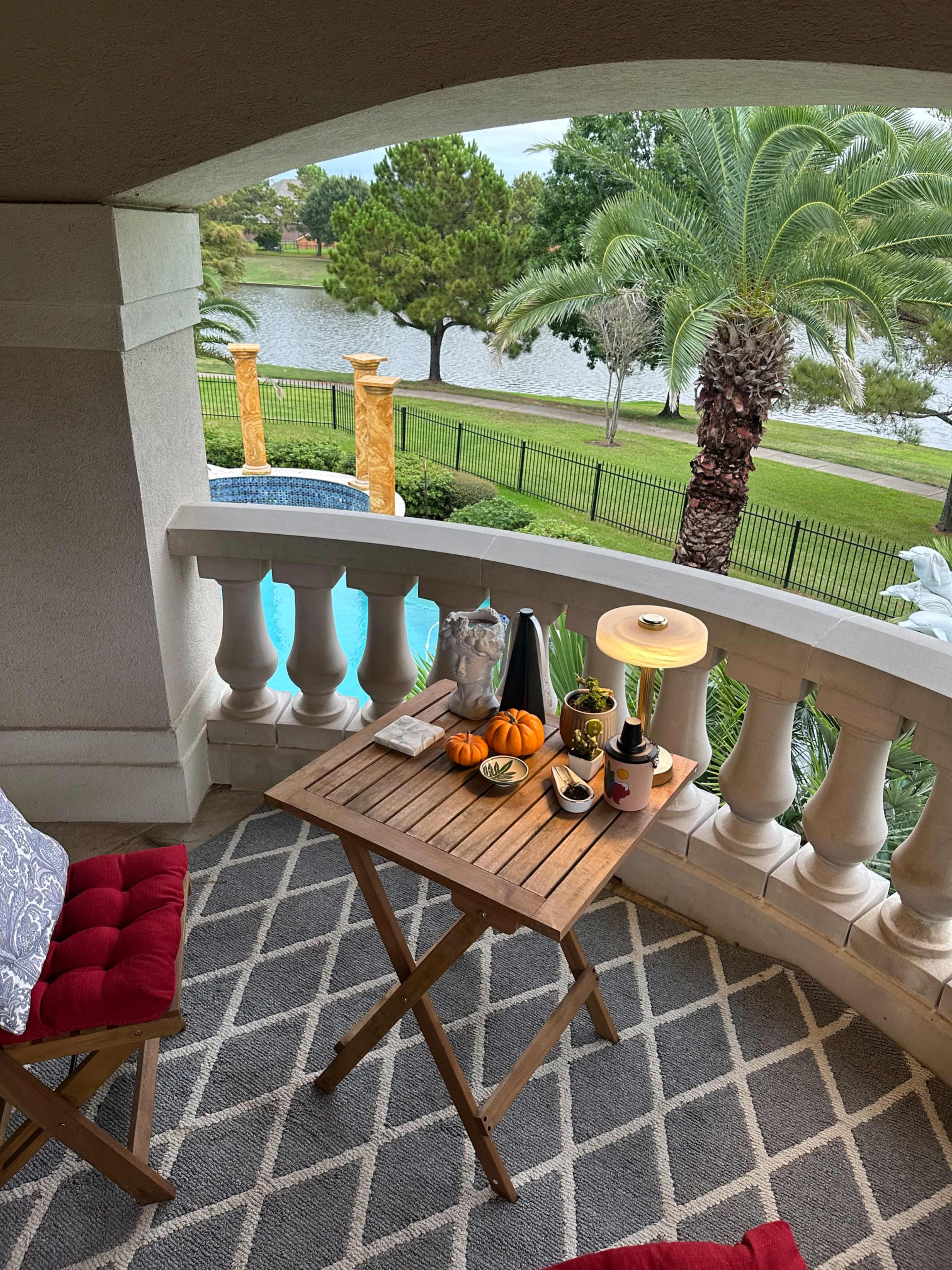 A small balcony features a wooden table with decorative pumpkins, a bottle of wine, and a lamp, overlooking a pool and manicured landscaping.