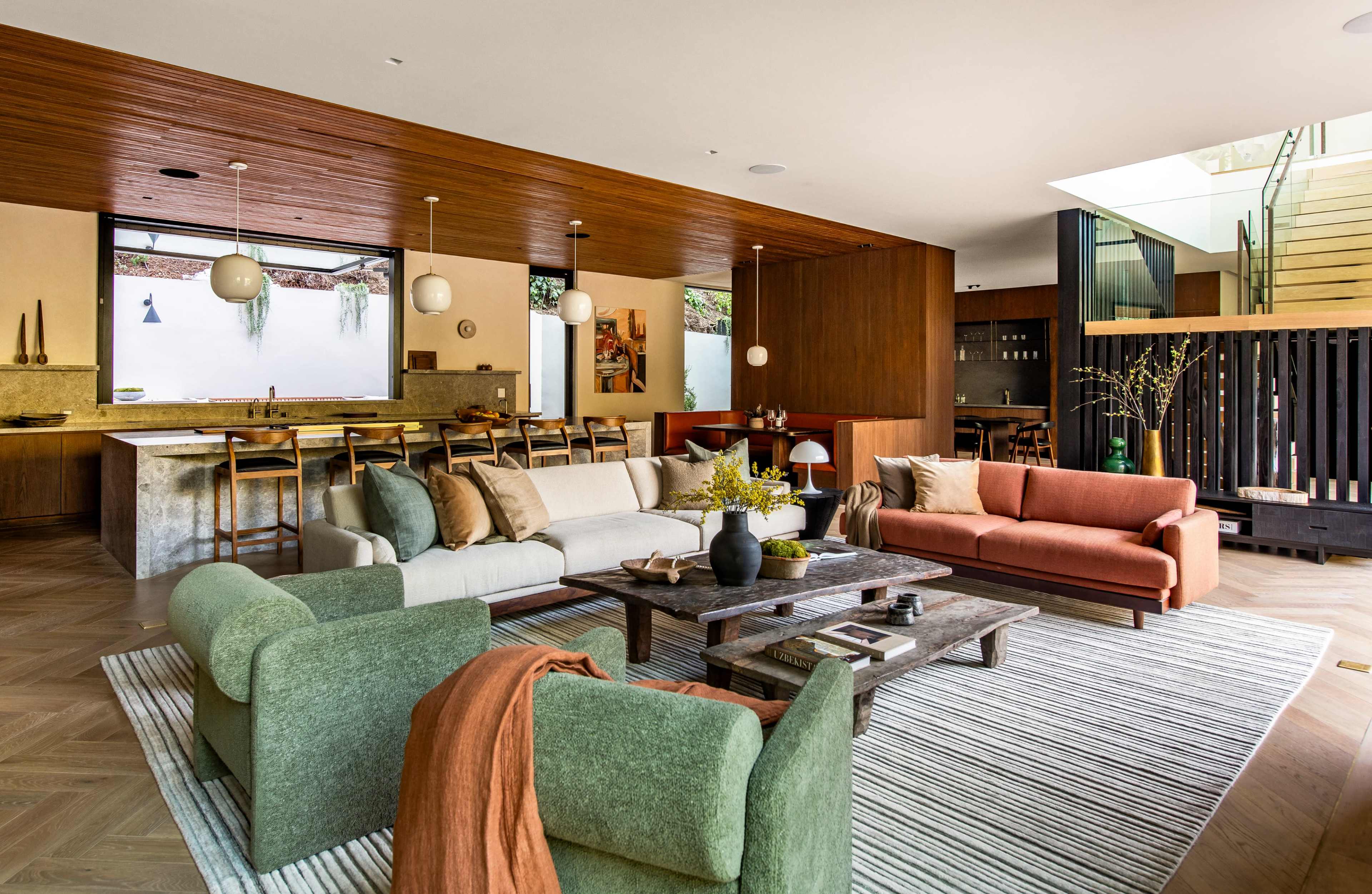 The image shows a modern living room with a mix of green, beige, and orange sofas, a wooden coffee table, and large windows that provide natural light.