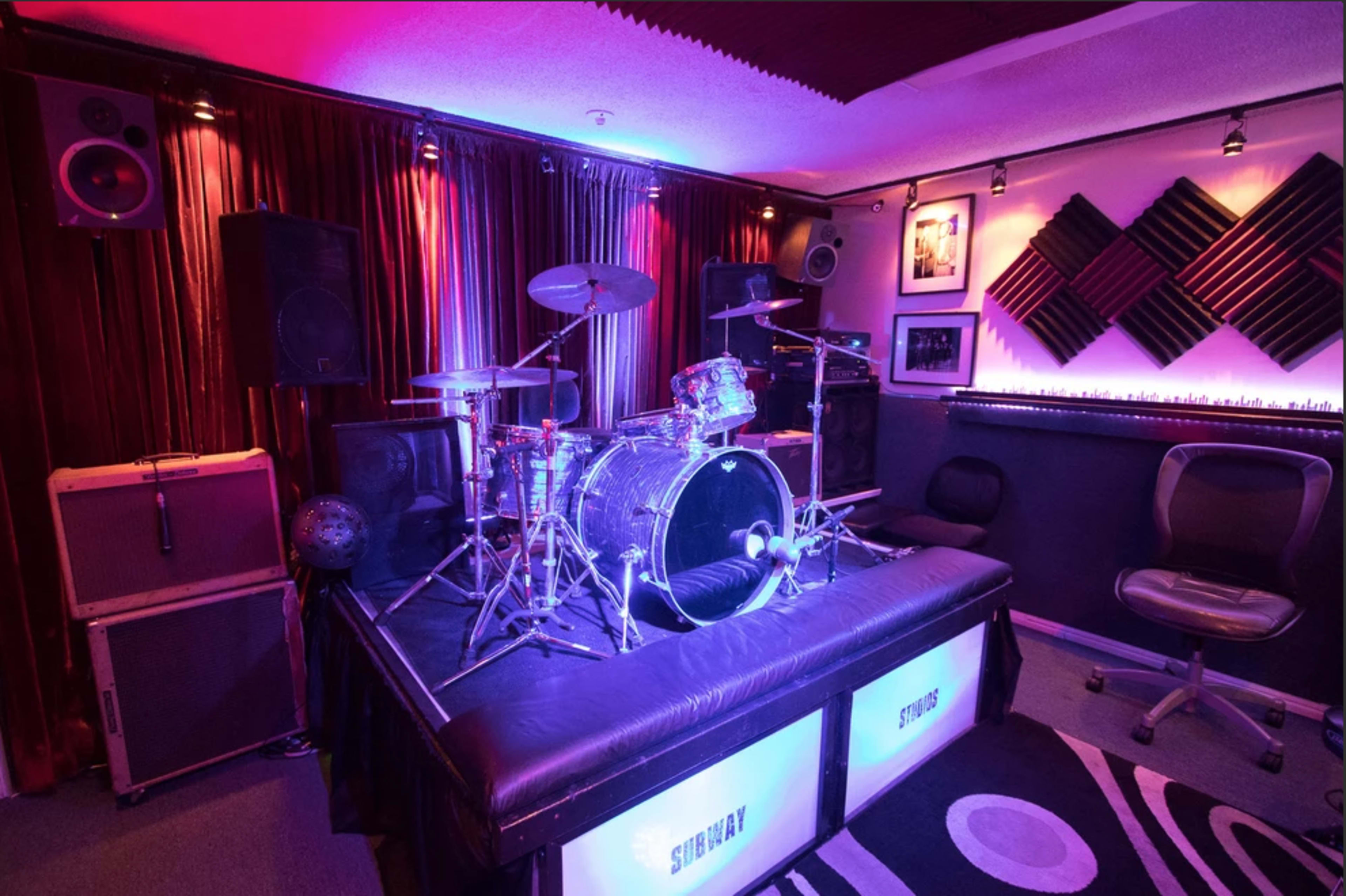 The image features a drum set with colorful lighting, surrounded by amplifiers, acoustic panels, and a lounge chair in a music studio setting.