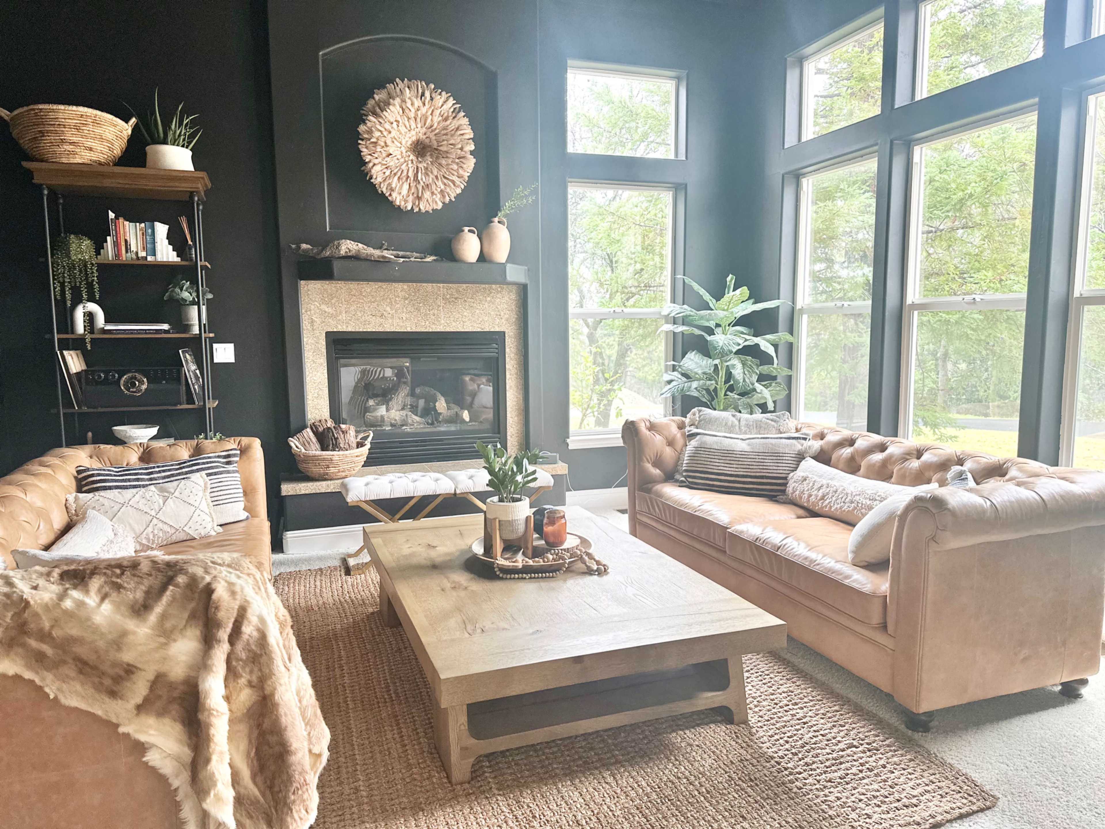 The living room features two leather chesterfield sofas facing a central wooden coffee table, with a fireplace and large windows providing a view of the outdoors.