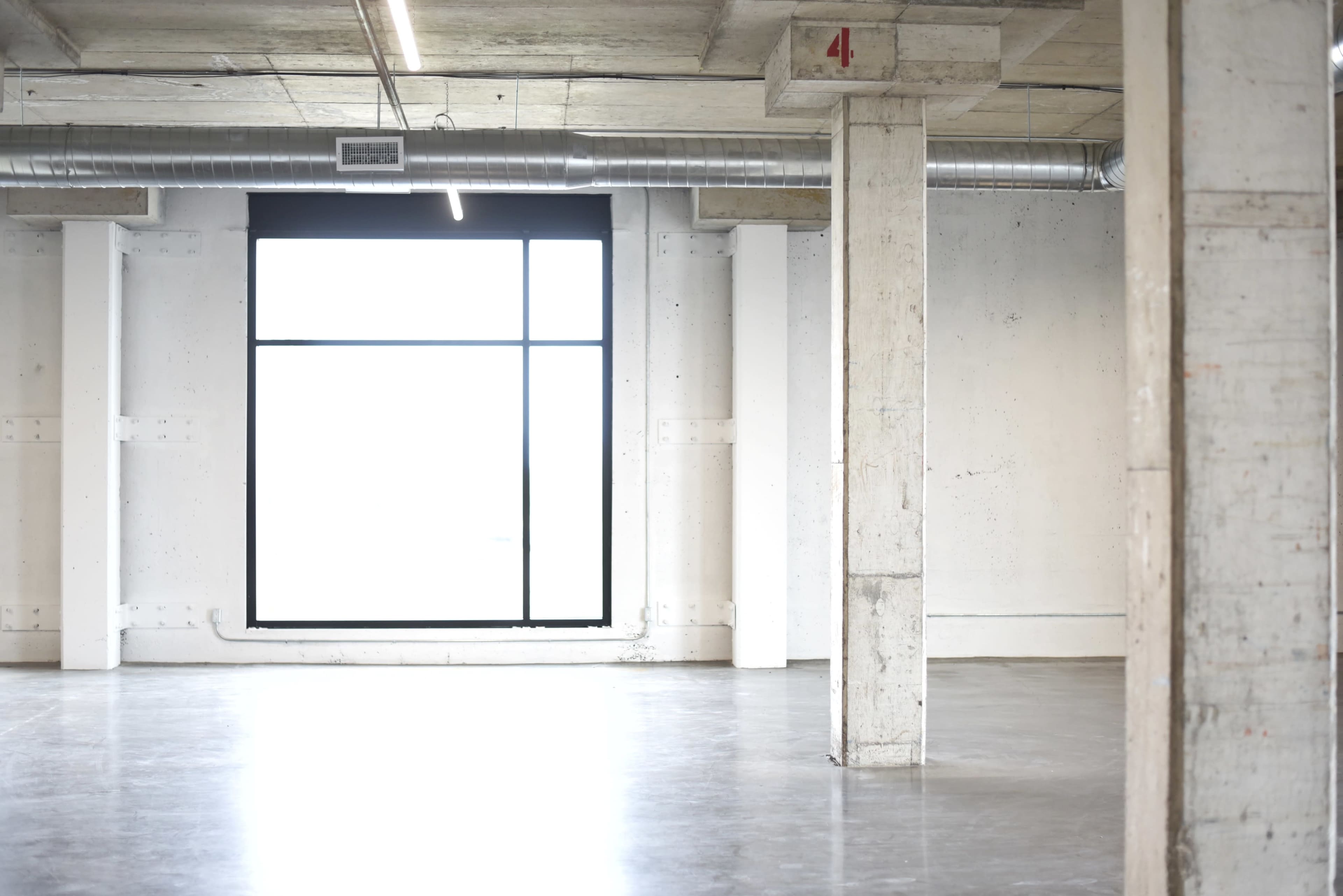 The image shows an empty industrial space with concrete walls, polished floors, and a large window casting light into the room.