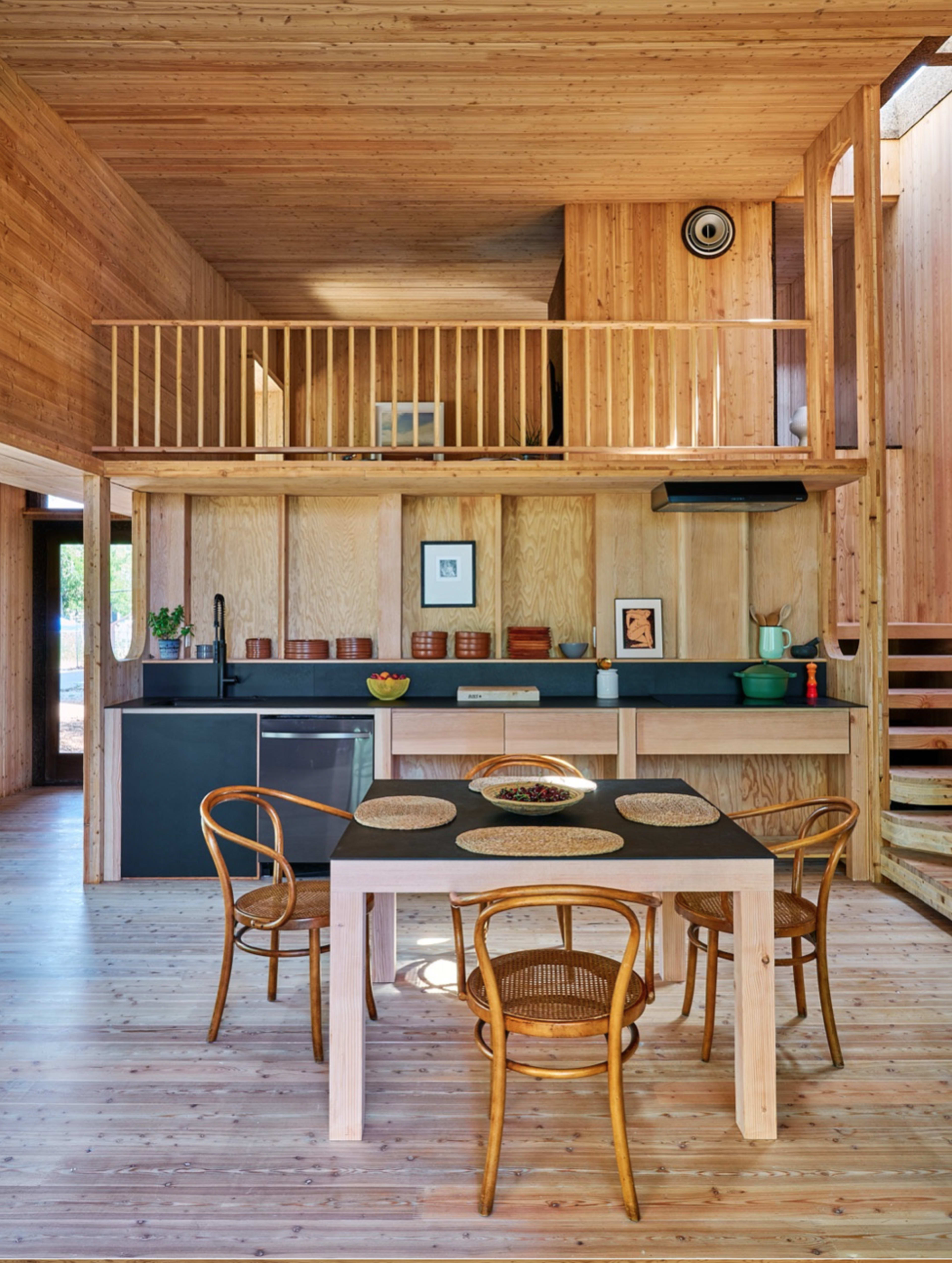 A wooden interior of a minimalist kitchen and dining area, featuring a table with chairs and a staircase leading to an upper loft.