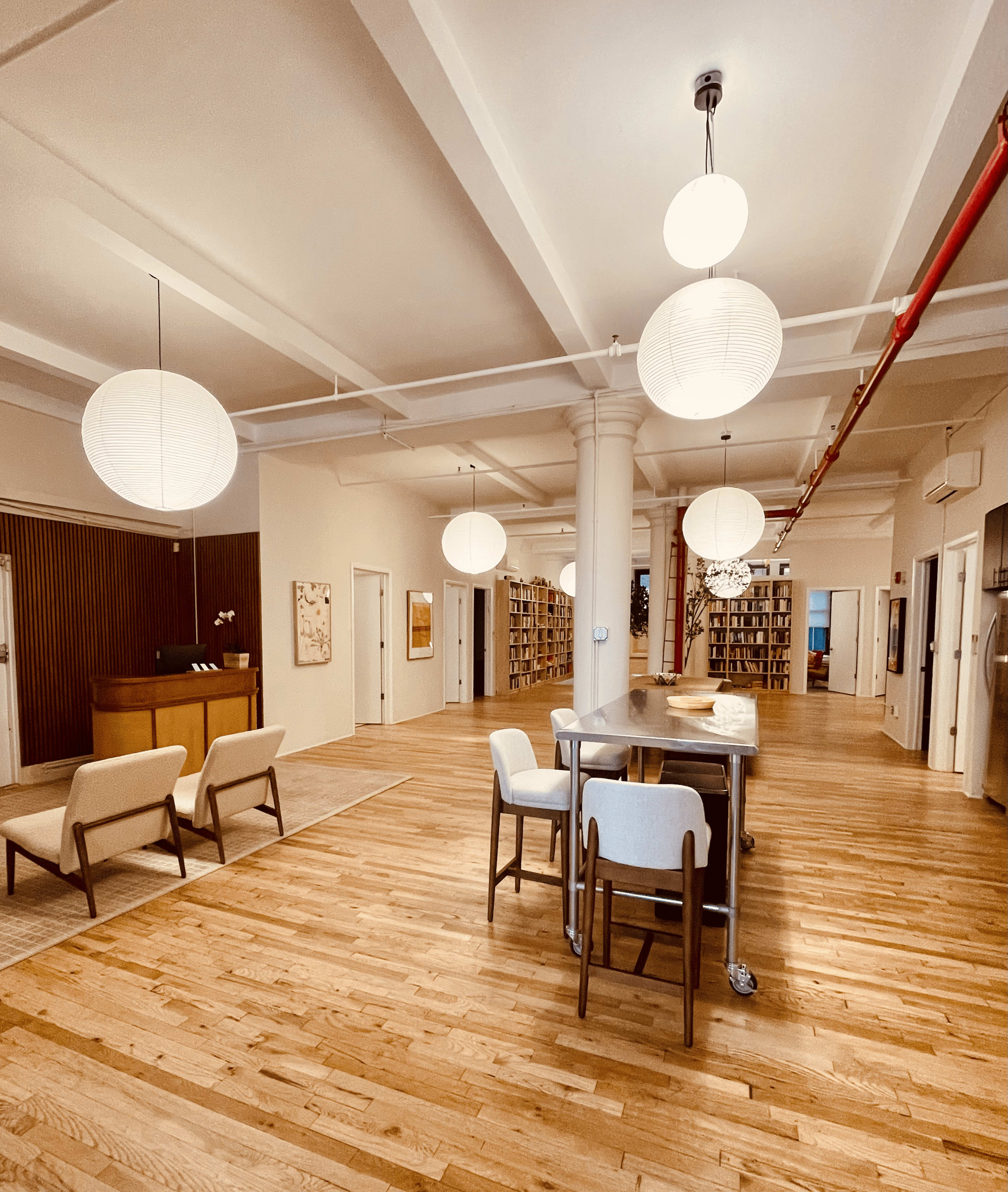 The image shows a spacious, modern office area with wooden flooring, round pendant lights, and a reception desk in the background surrounded by bookshelves.