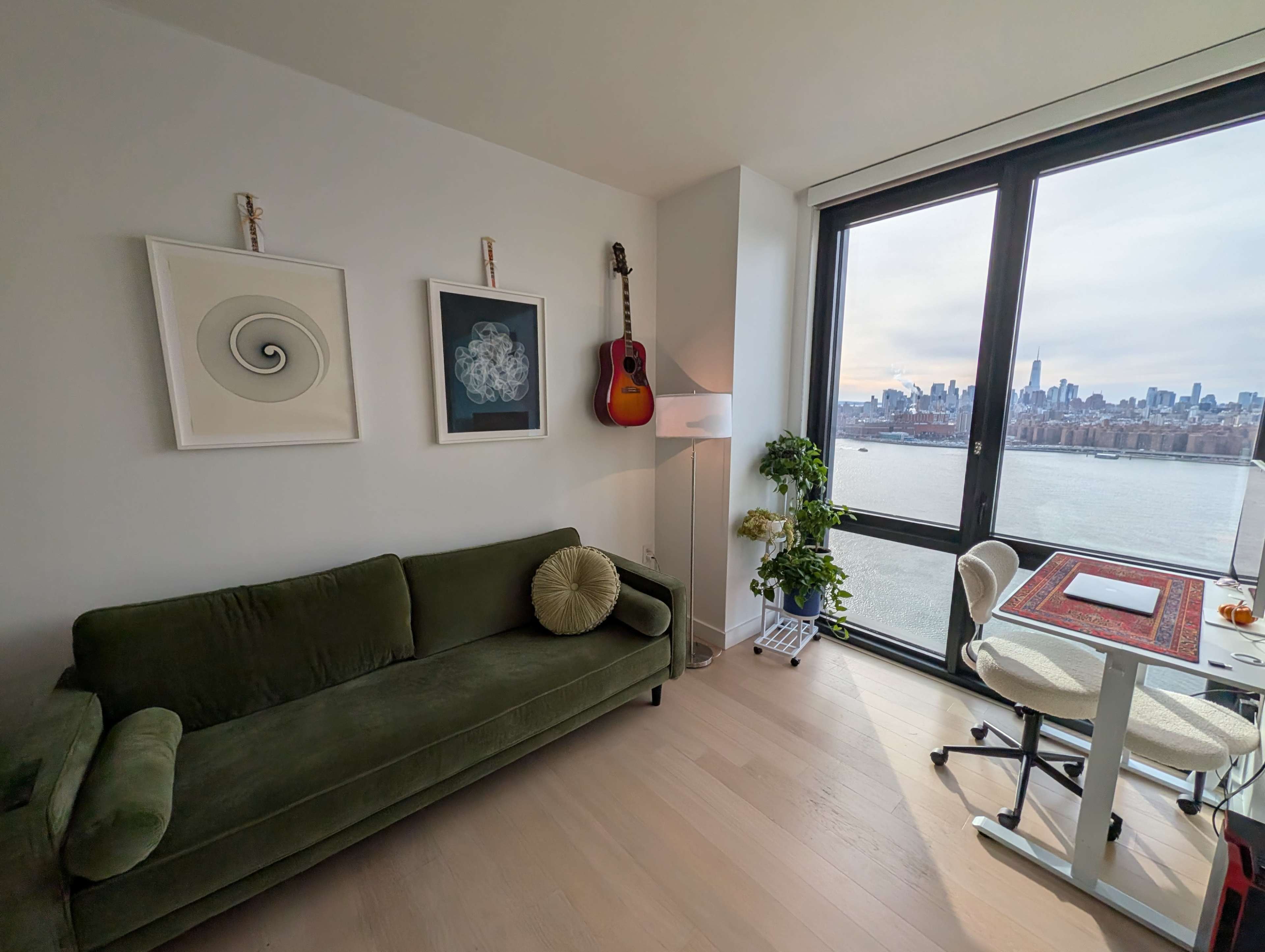 A modern living space features a green sofa, a framed artwork, a guitar mounted on the wall, a small plant, and a desk with a view of the city skyline through large windows.