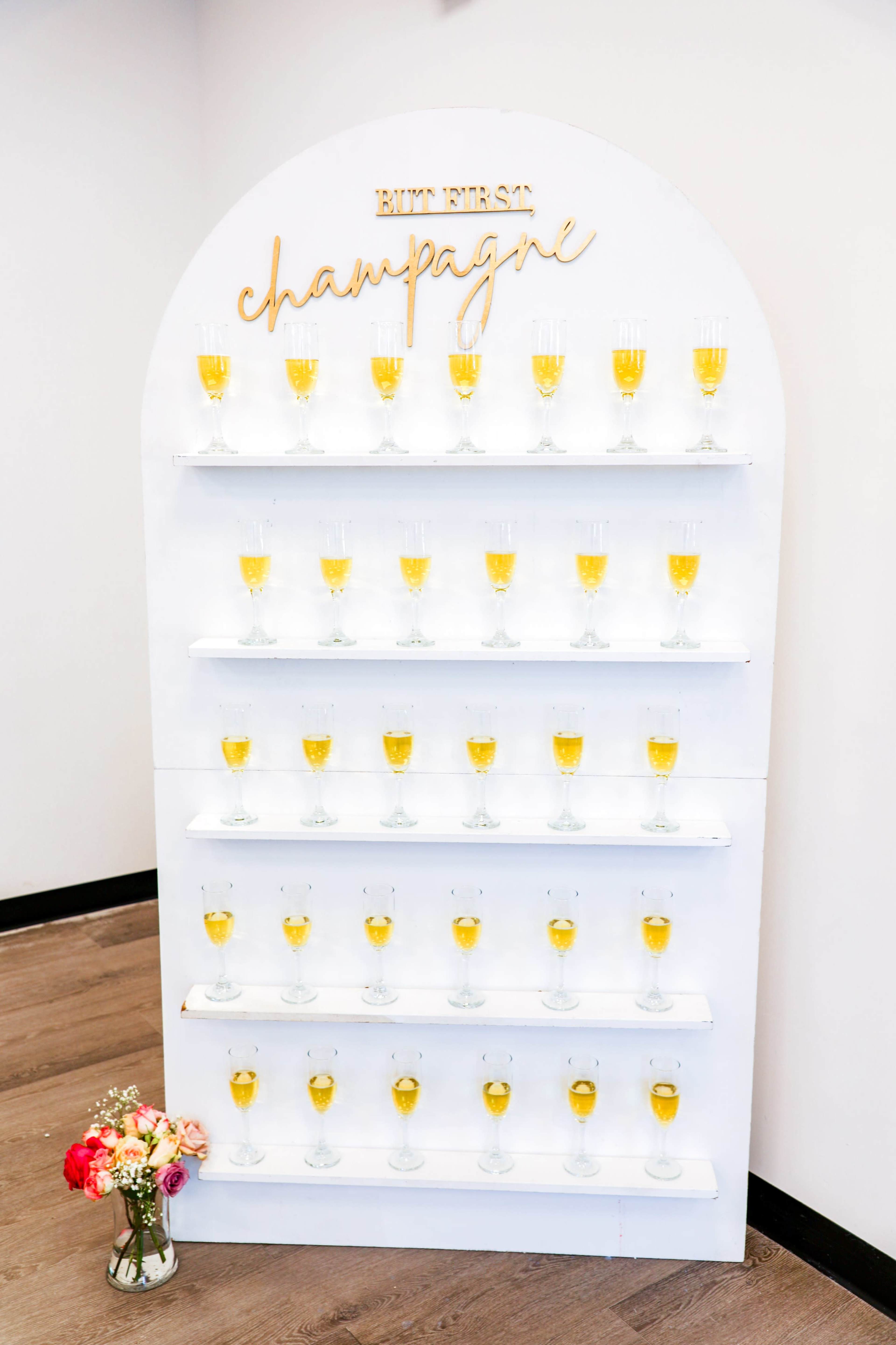 A decorative wall display features multiple rows of champagne glasses filled with a golden drink, accompanied by the phrase "BUT FIRST, champagne" at the top.