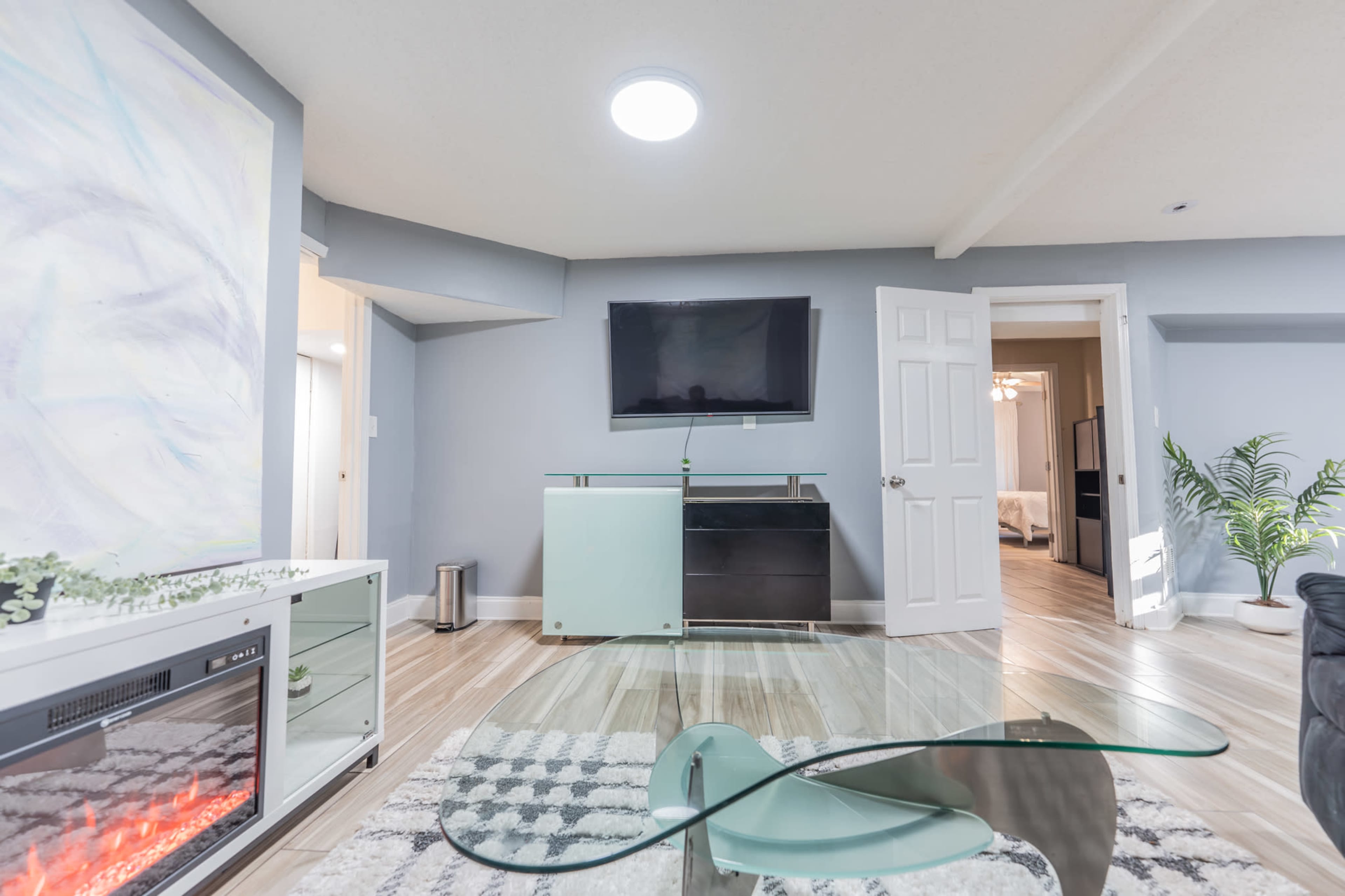 The image shows a modern living room with a television mounted on the wall, a glass coffee table, and a fireplace, along with a view of an adjacent room.