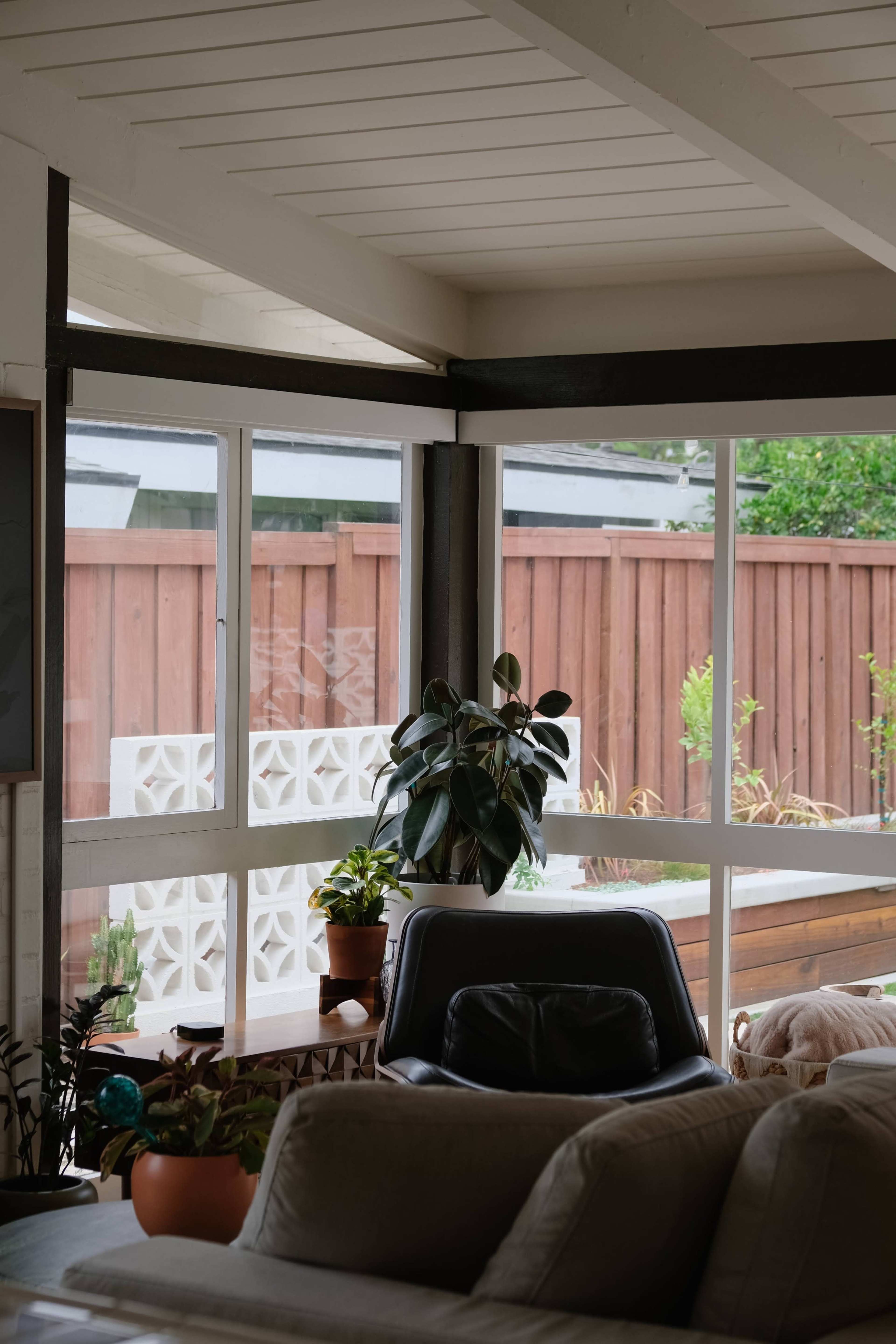 The image shows a modern living room with large windows overlooking a wooden fence and greenery, featuring a cozy couch and potted plants.