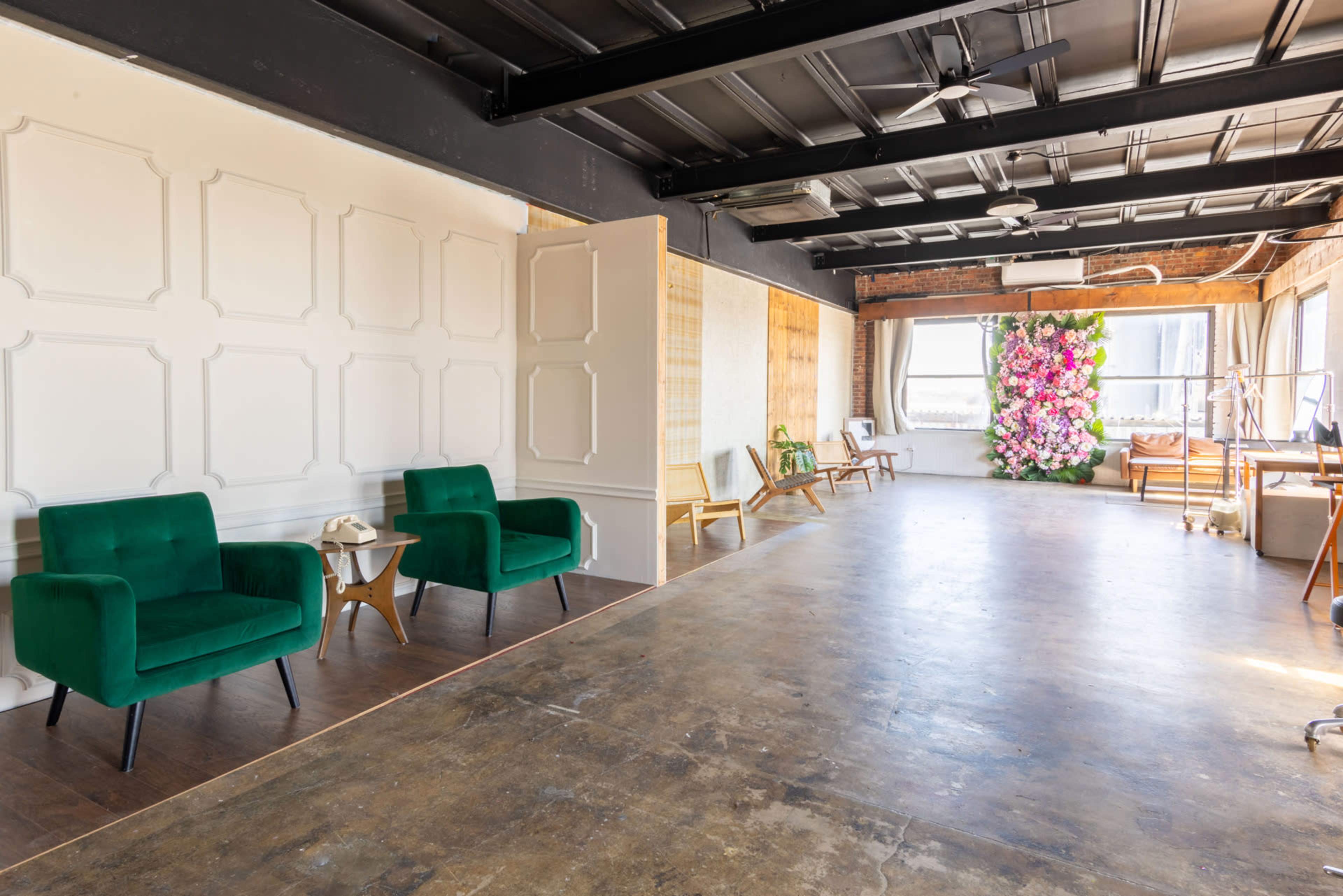 The interior of a spacious, well-lit room features two green chairs, a small side table, and a floral arrangement against a wall, with industrial-style ceilings and large windows.