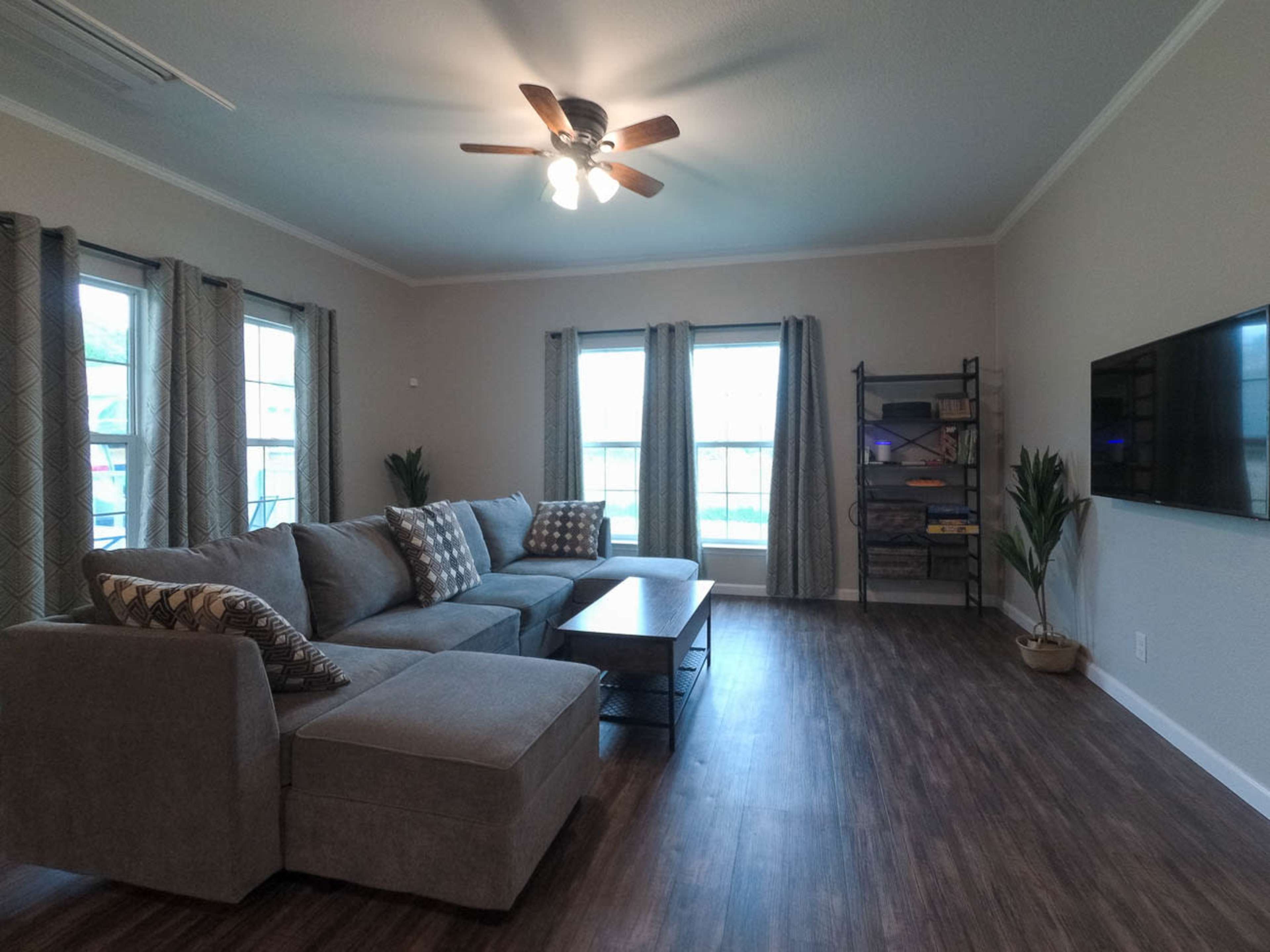 The living room features a sectional sofa with decorative pillows, a coffee table, a TV mounted on the wall, and large windows with curtains.
