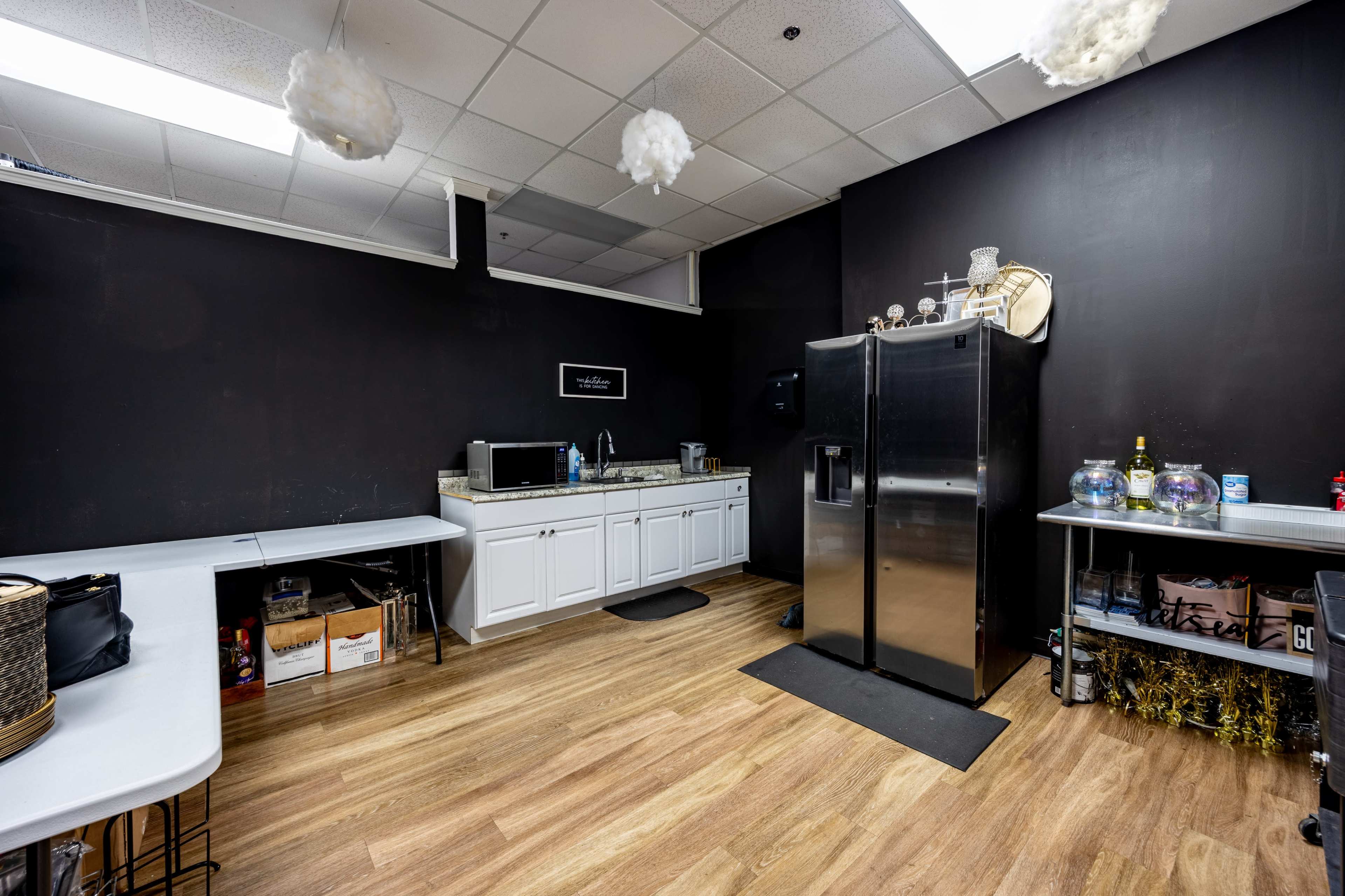 The image shows a small kitchenette area with a black wall, a counter with cabinets, a large refrigerator, and several tables.