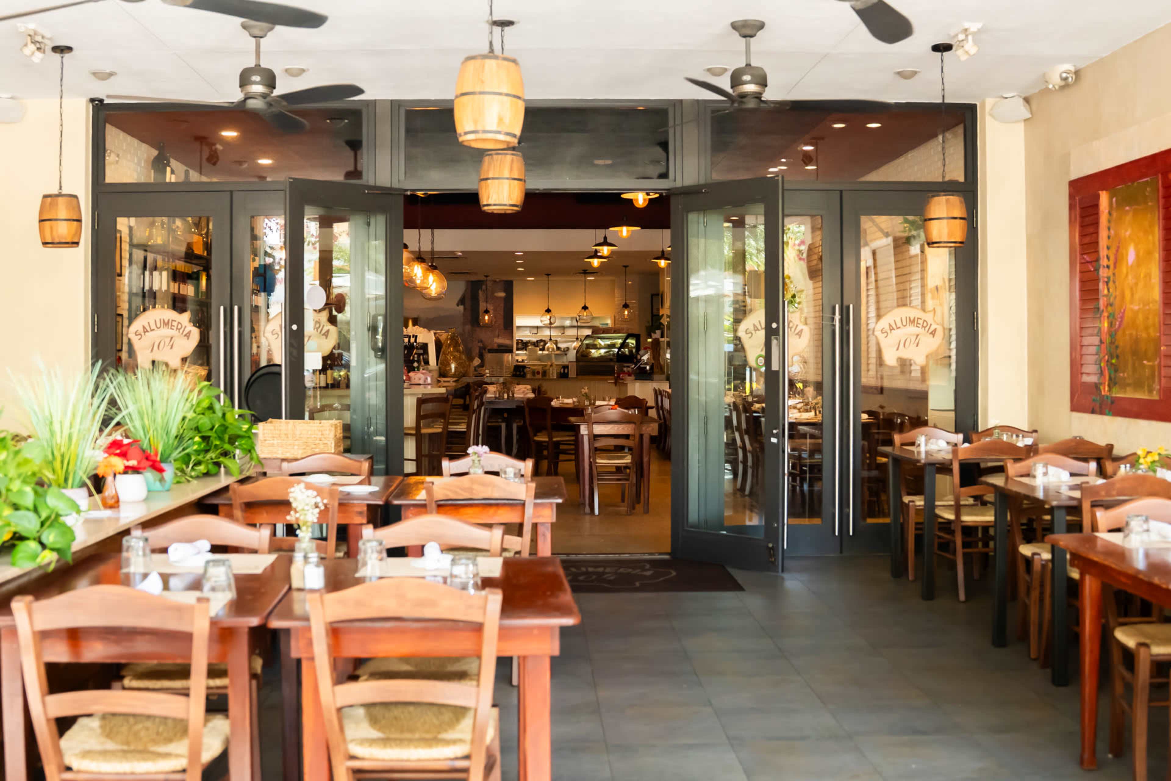The image shows a restaurant entrance with open double doors leading to a warmly lit dining area, featuring wooden tables and chairs arranged both inside and outside.