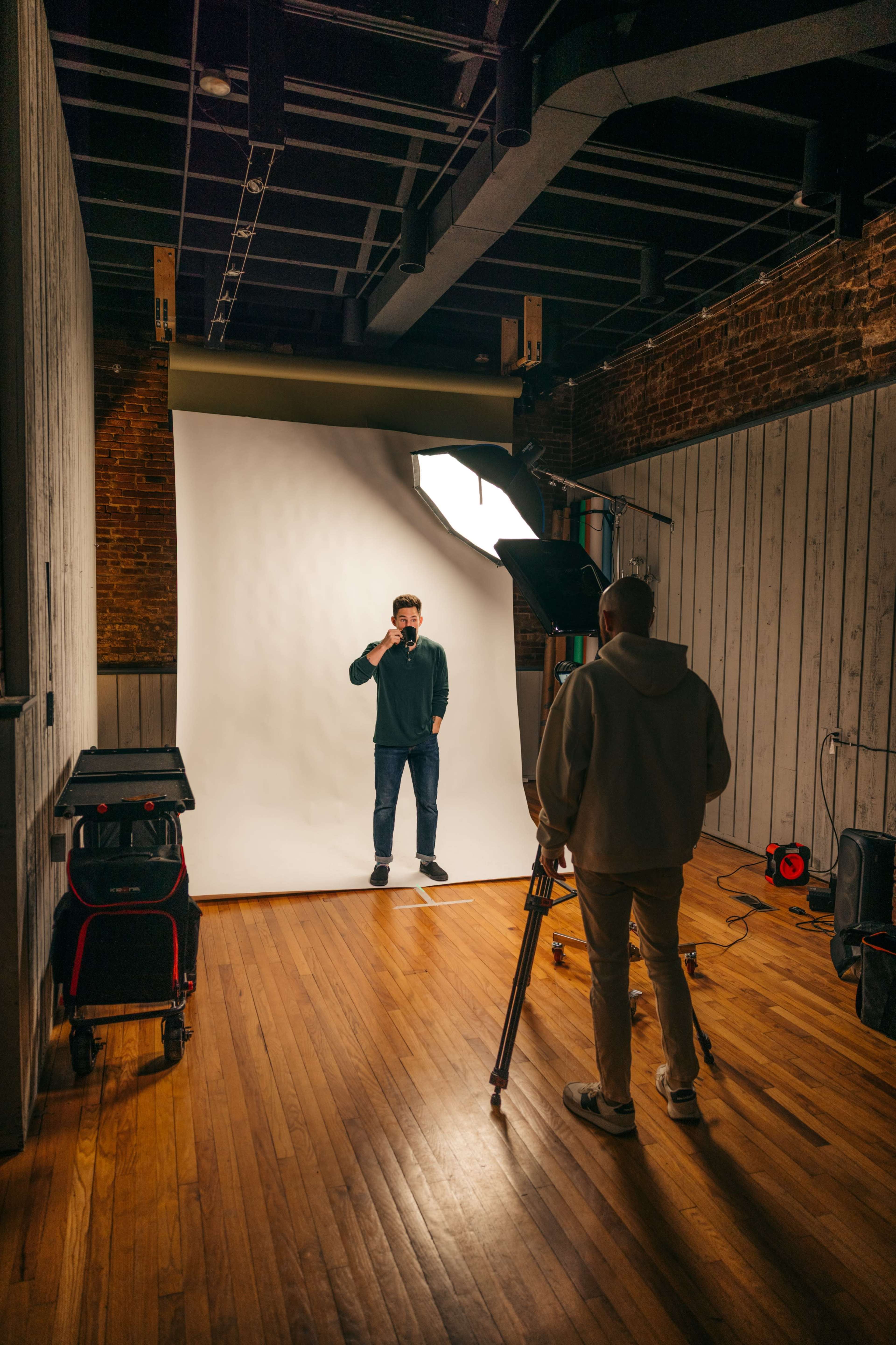 A man stands in front of a light backdrop while another man operates a camera on a tripod in a studio setting.