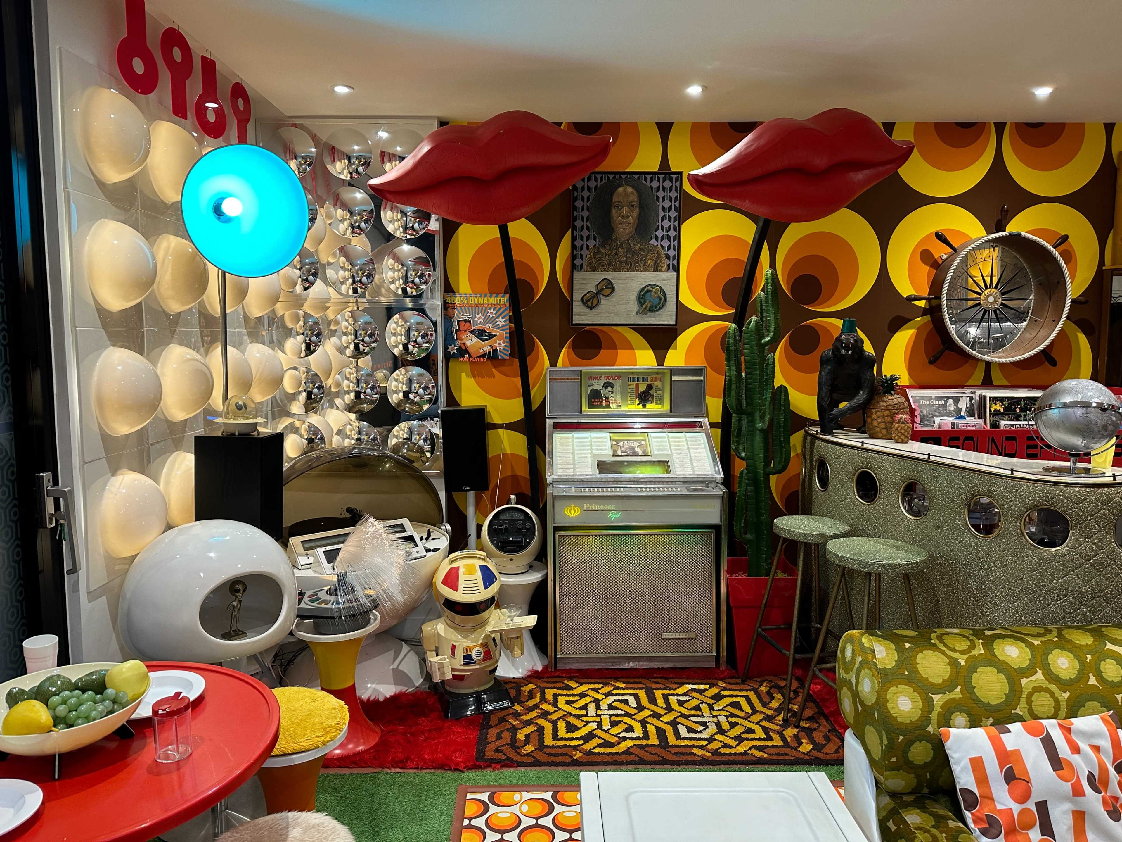 A vibrant retro-themed room filled with colorful furniture, vintage decor, and unique art pieces, including large lip-shaped structures and a vintage jukebox.
