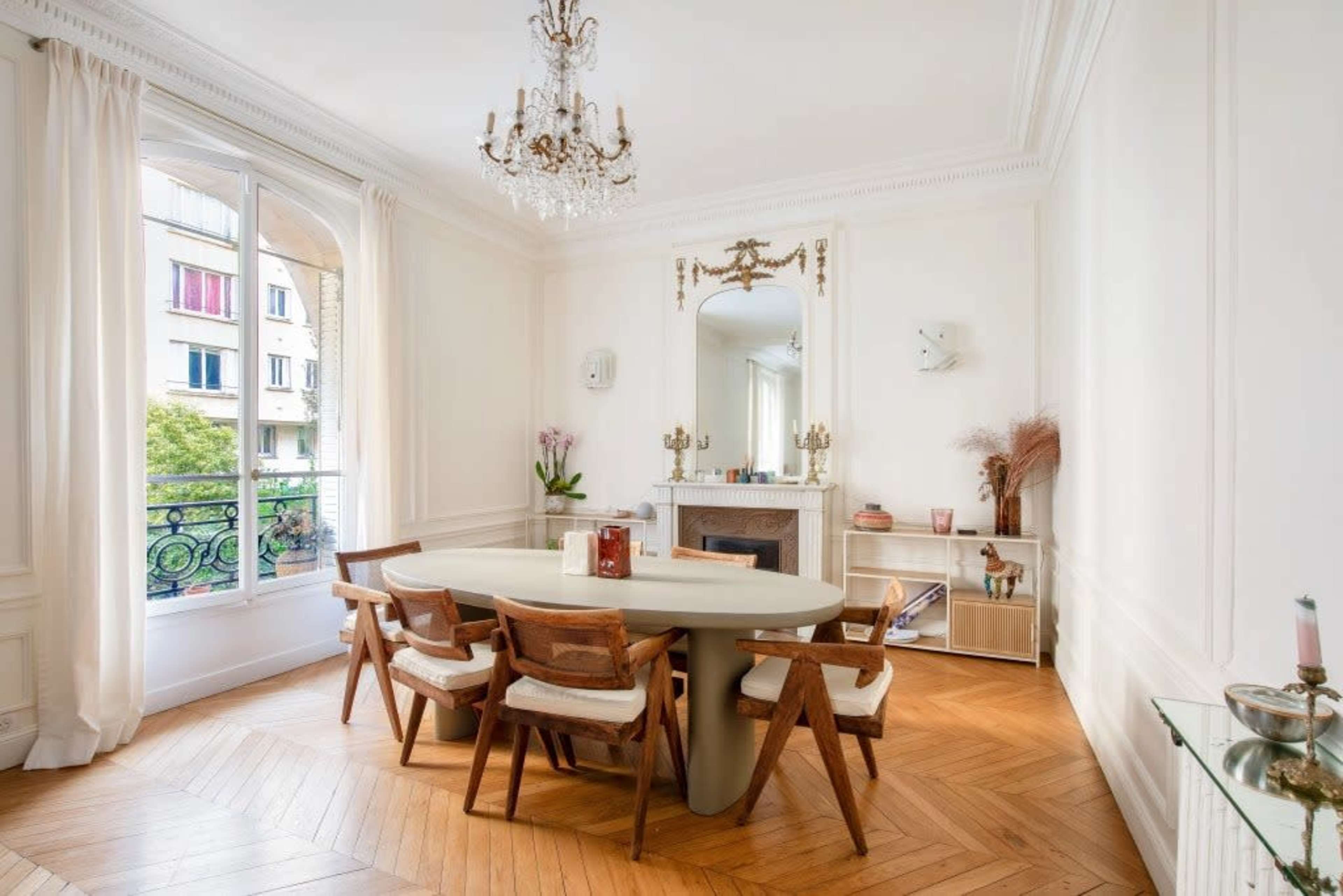 The image shows a bright dining room with a round table, wooden chairs, and a decorative fireplace, complemented by large windows and light-colored walls.