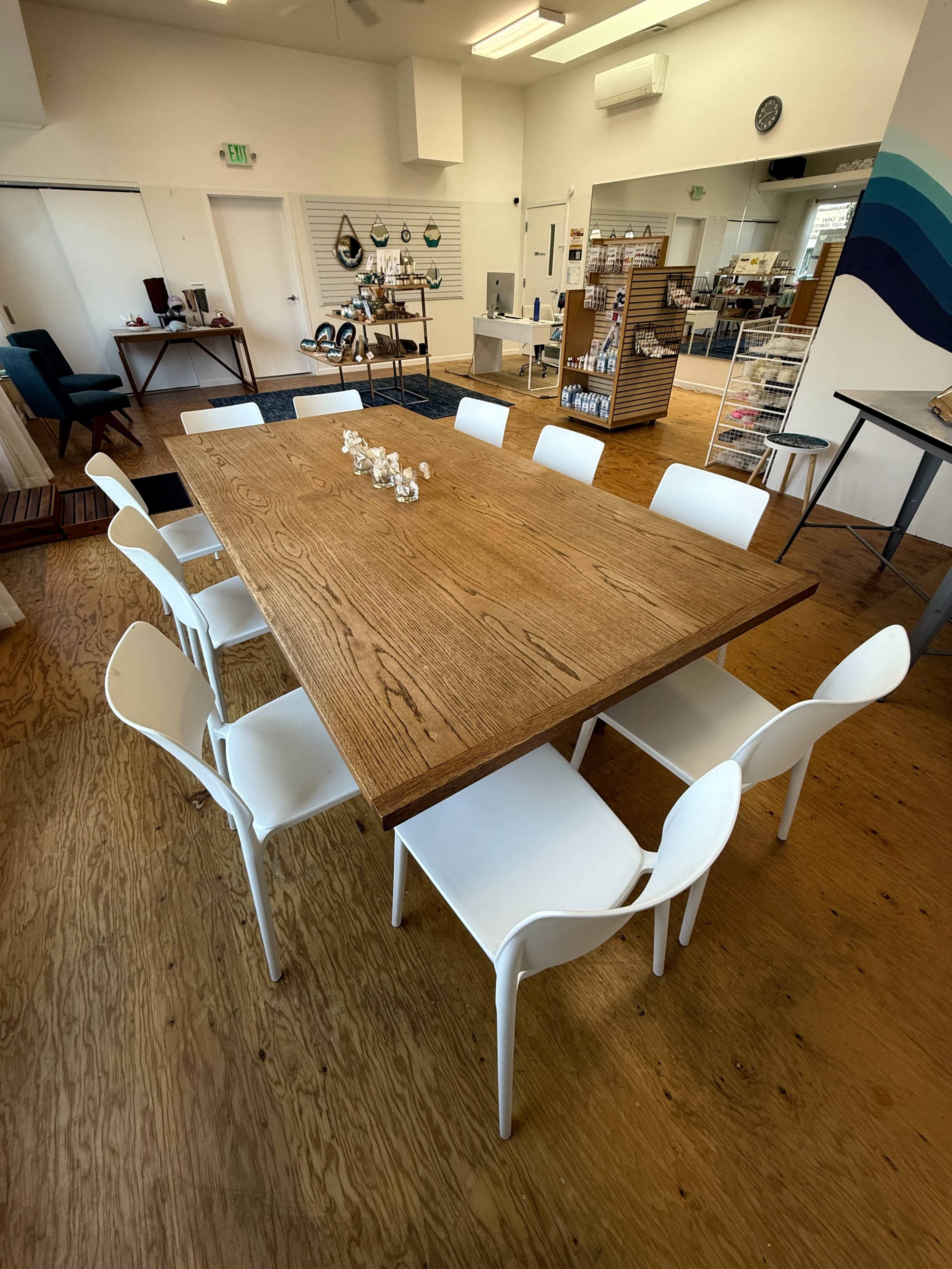 A spacious room features a large wooden table surrounded by white chairs, with various display shelves and products in the background.