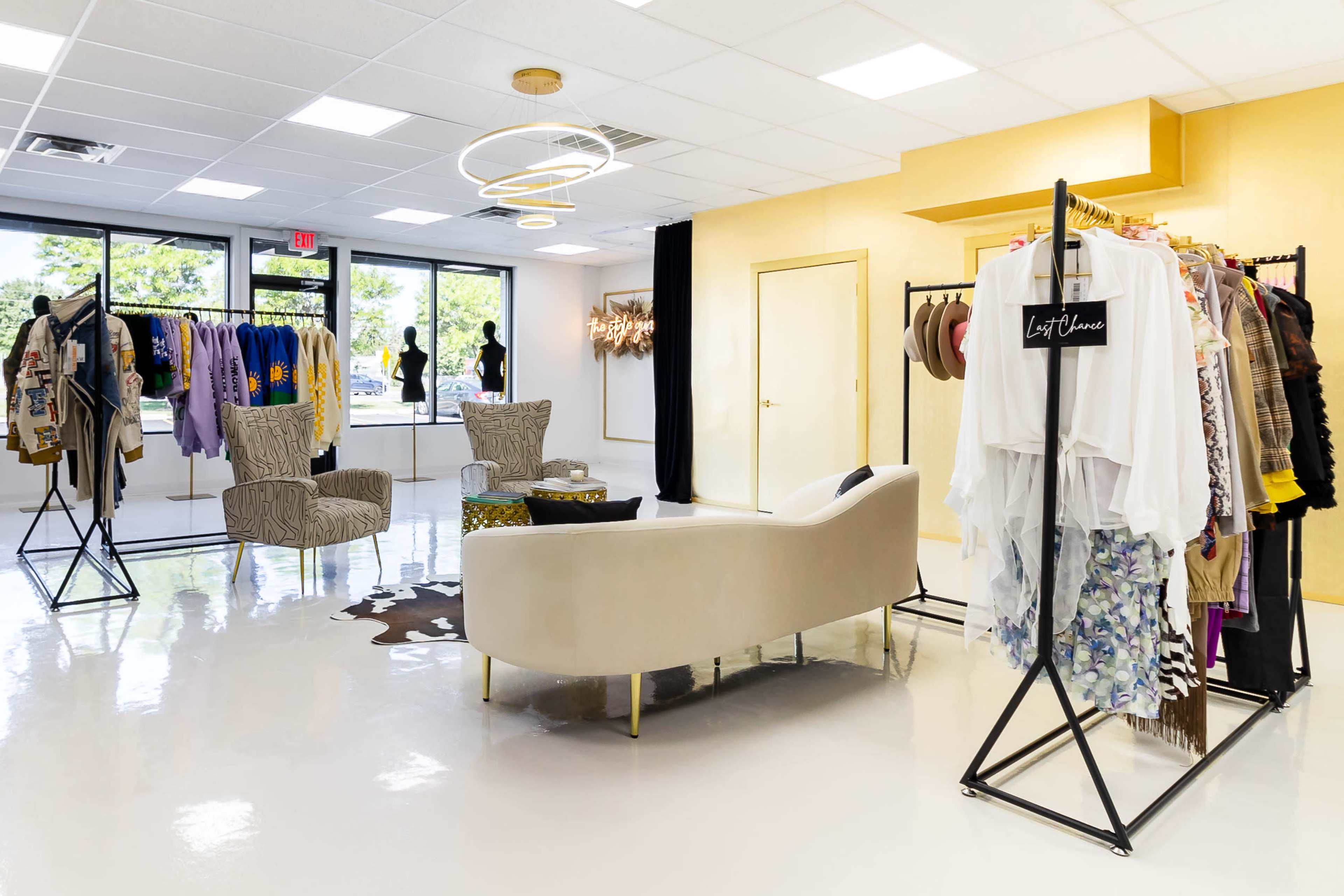 A stylish boutique interior featuring clothing racks, a seating area with two chairs, and large windows allowing natural light to enter.