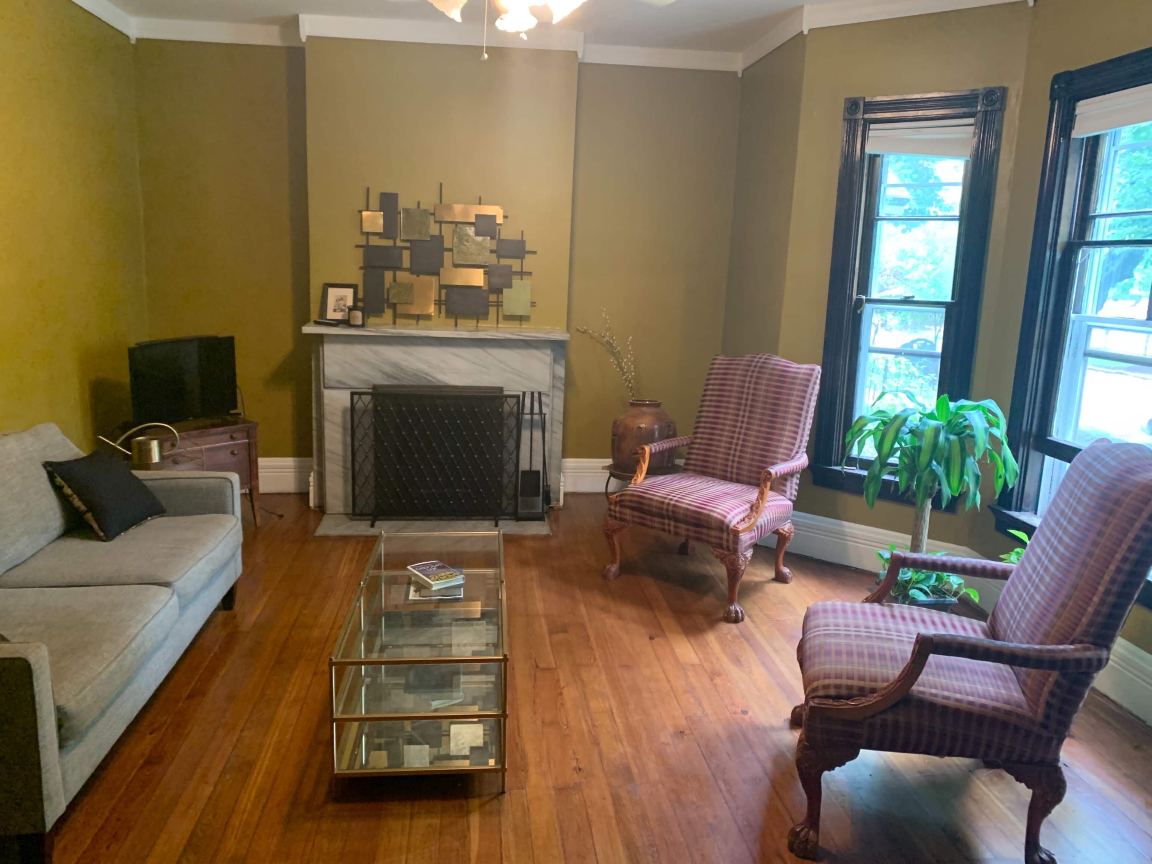 A living room with a gray sofa, two patterned armchairs, a glass coffee table, and a marble fireplace against a green wall.