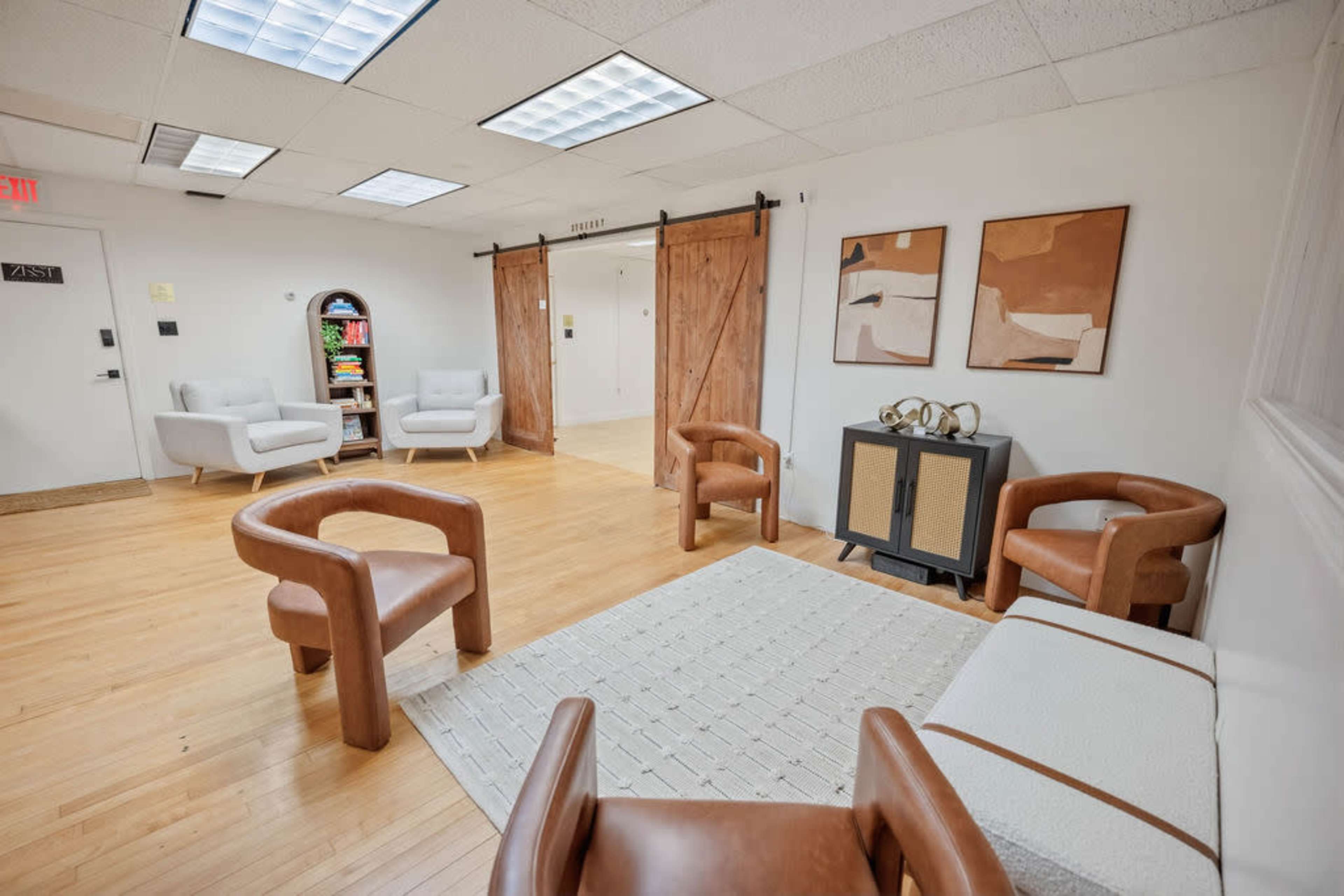 The image shows a spacious waiting room with wooden floors, three leather armchairs, a decorative side table, and two art pieces on the wall.