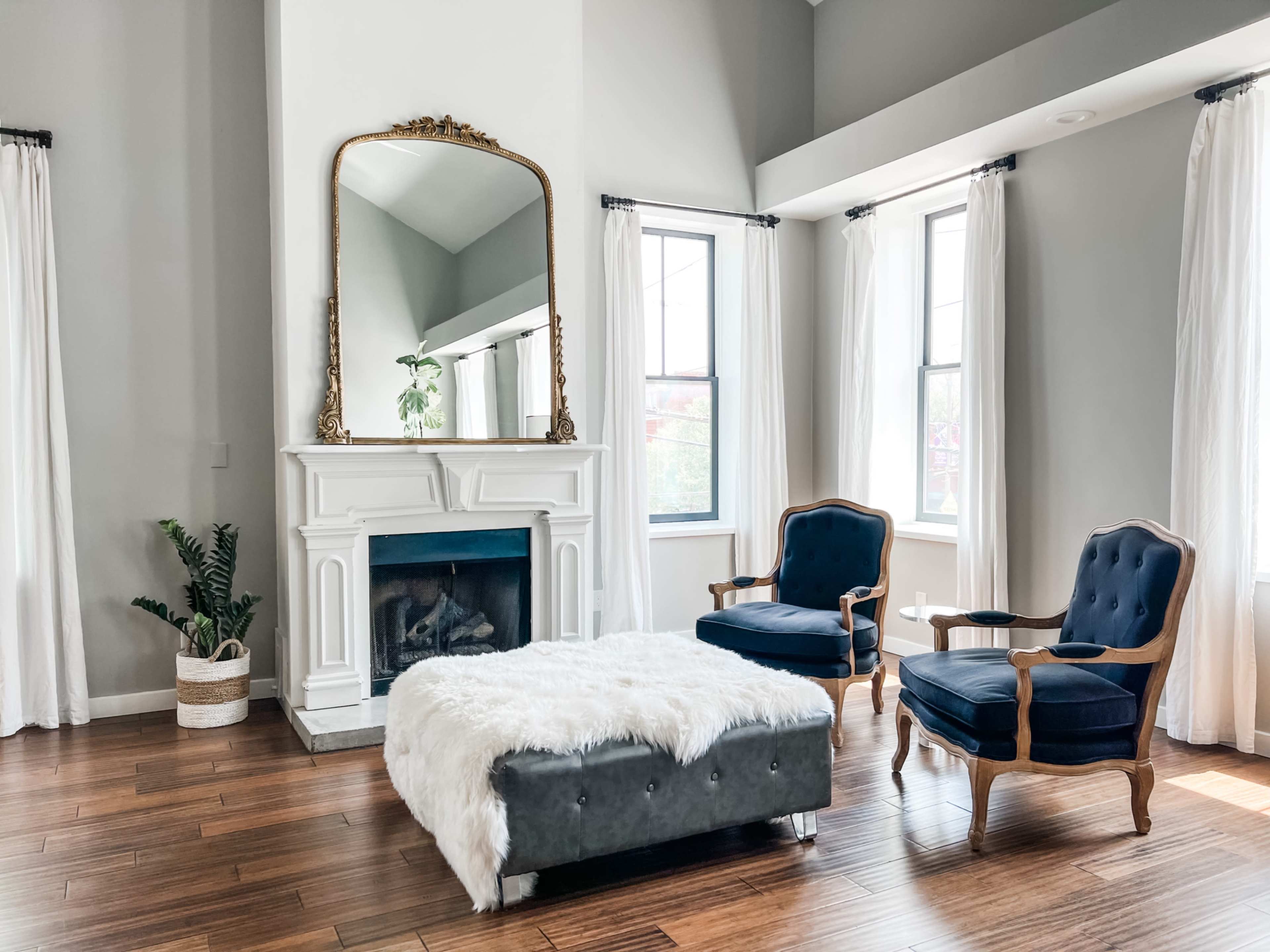 The image shows a modern living room featuring a fireplace with a large mirror above it, flanked by two blue chairs and a tufted ottoman on a hardwood floor.