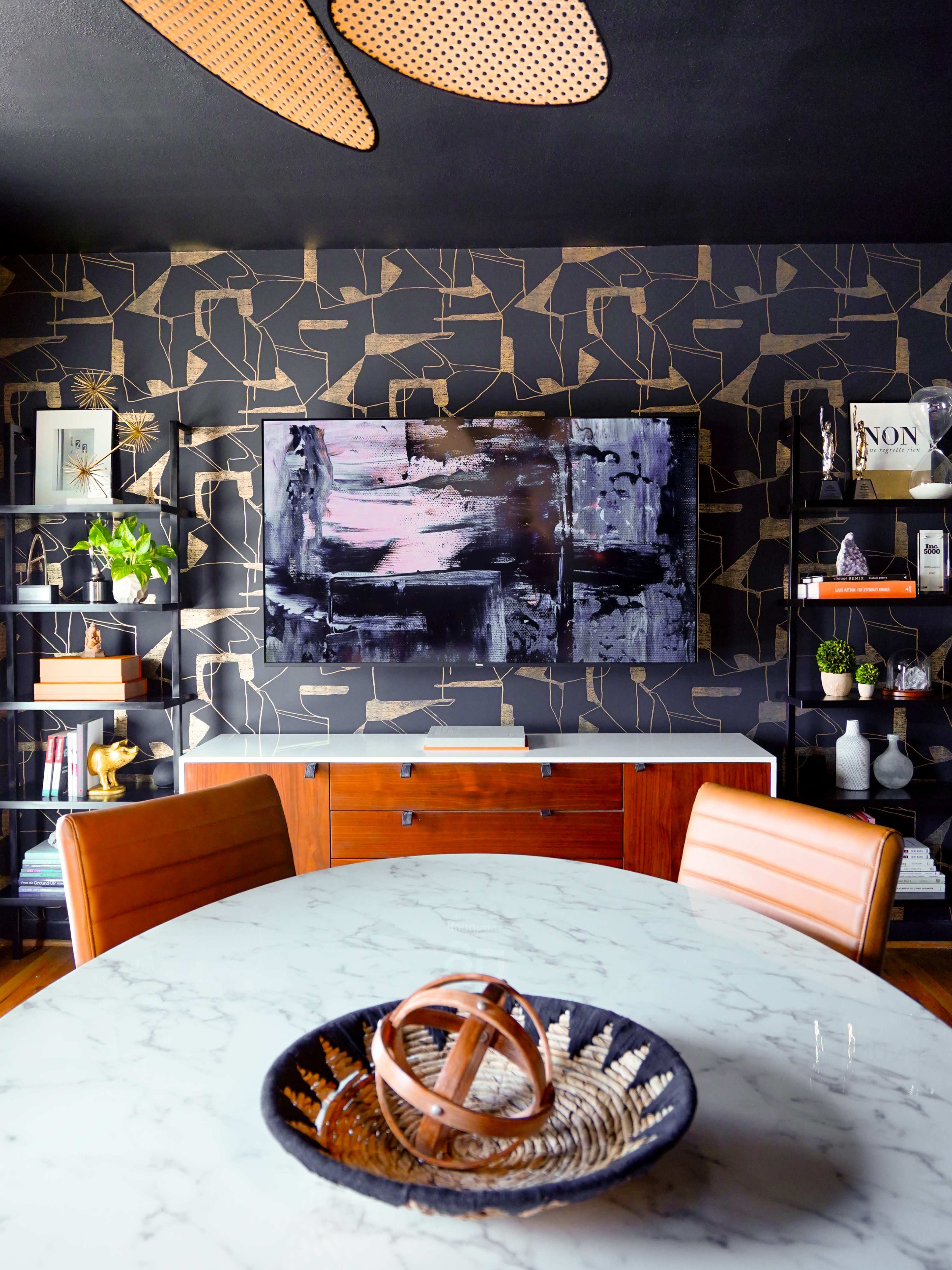 A modern dining area features a marble table, brown leather chairs, and a black wall with abstract art and shelves displaying decorative items.