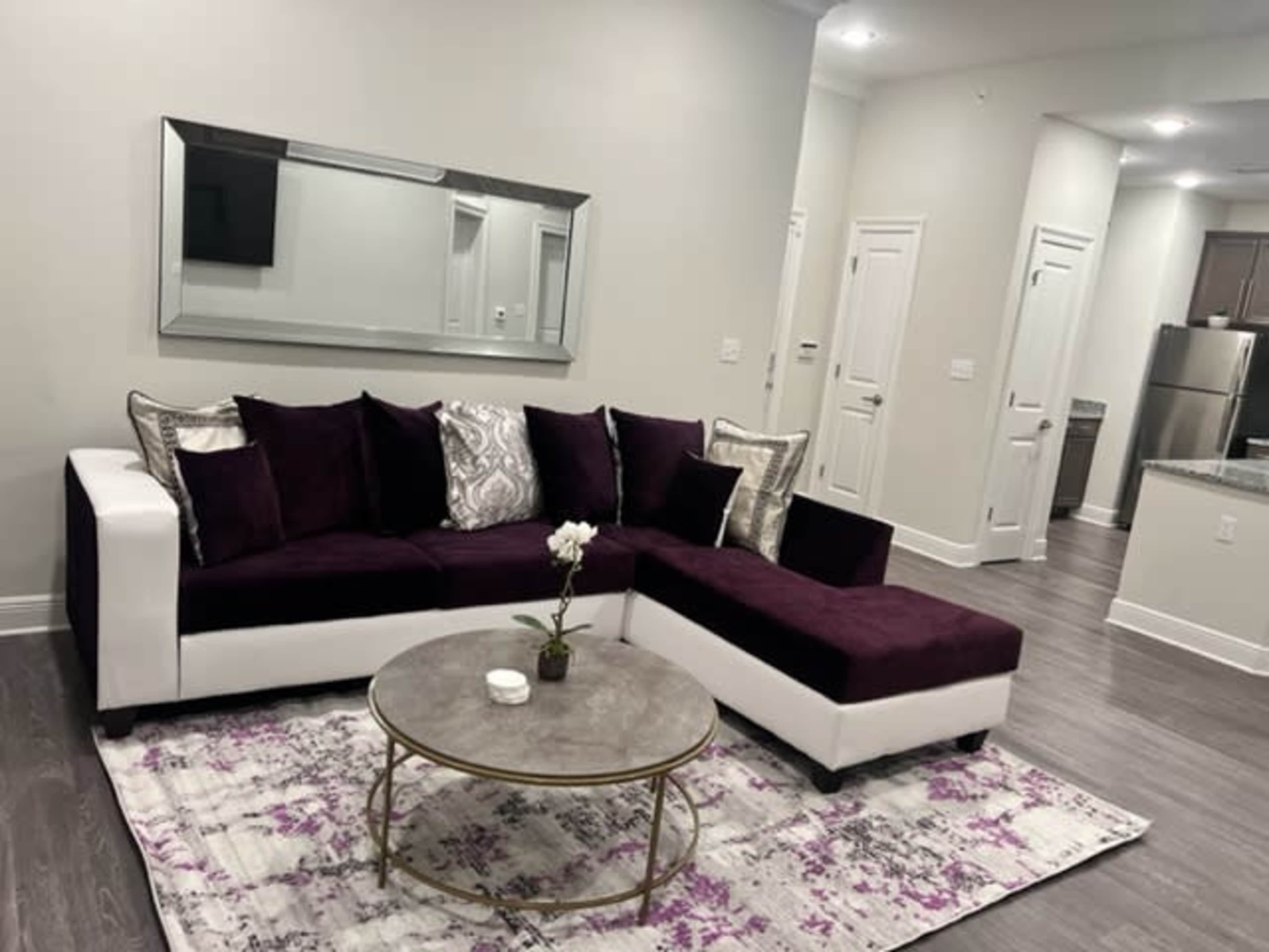 The image shows a modern living room featuring a purple and white sectional sofa, a round coffee table, and a mirror on the wall, with a kitchen visible in the background.