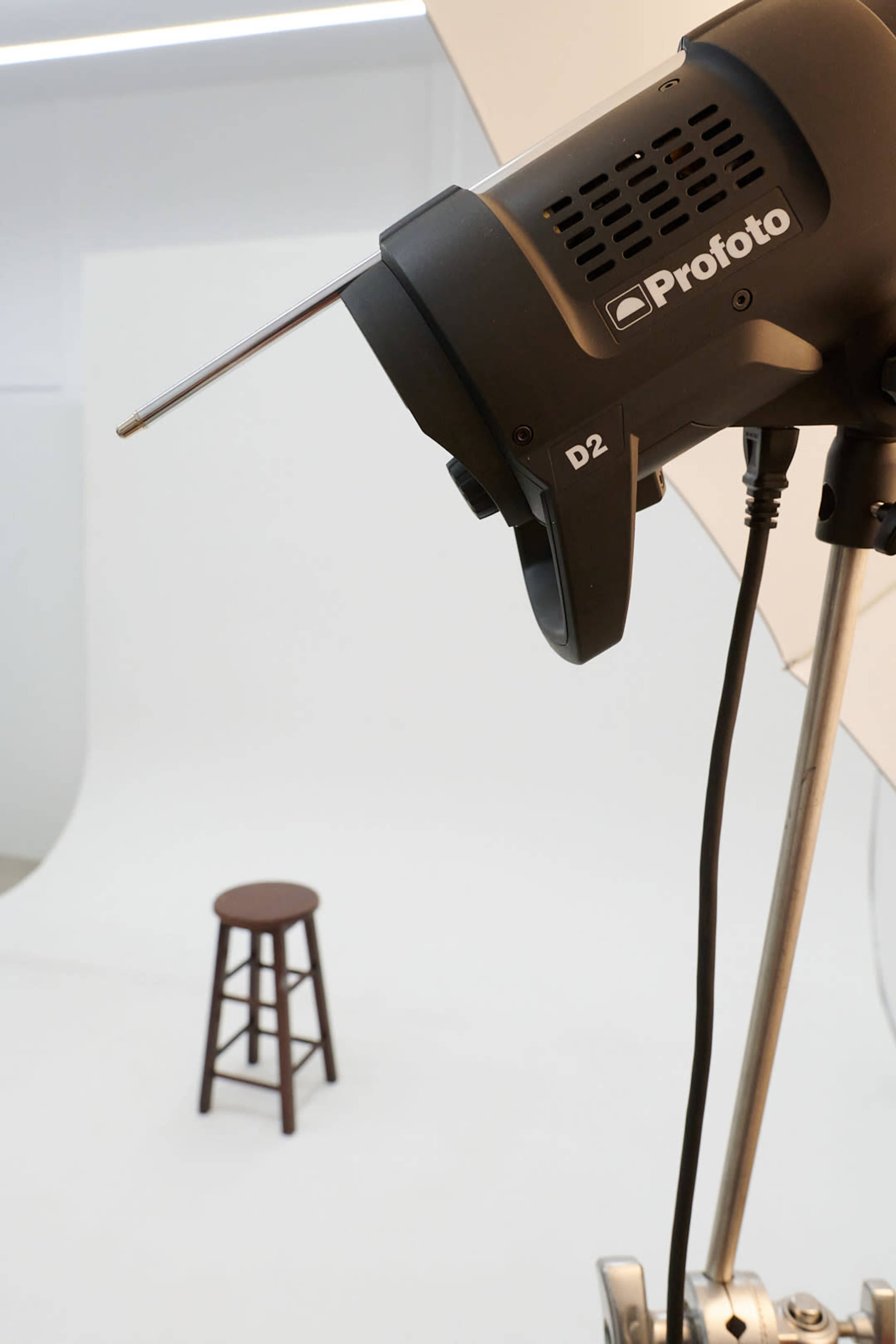 A Profoto D2 studio light is positioned above a wooden stool in a white photo backdrop setting.