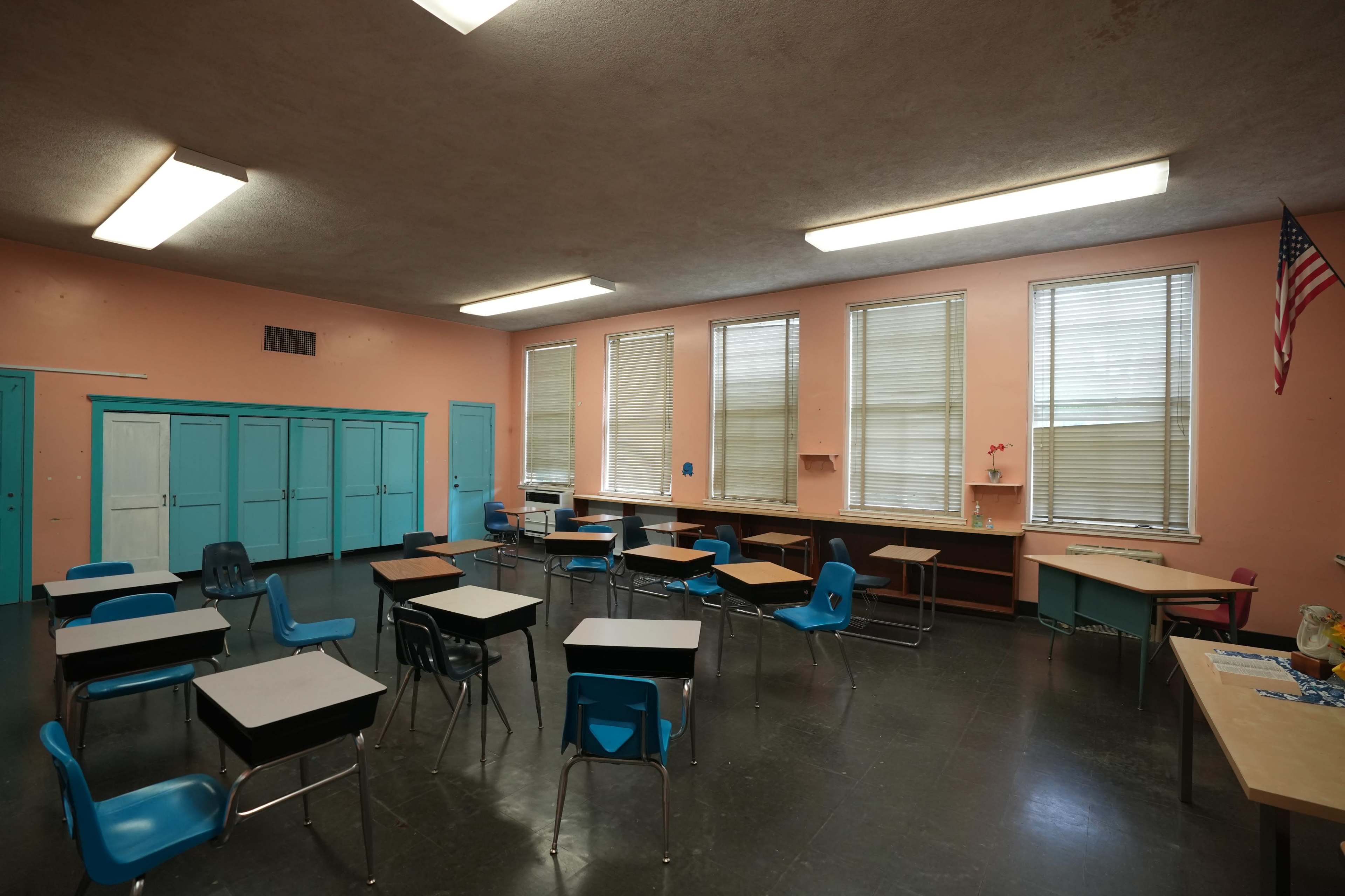 A classroom features several empty desks and chairs, with large windows and pink walls, along with cabinets and an American flag in the corner.