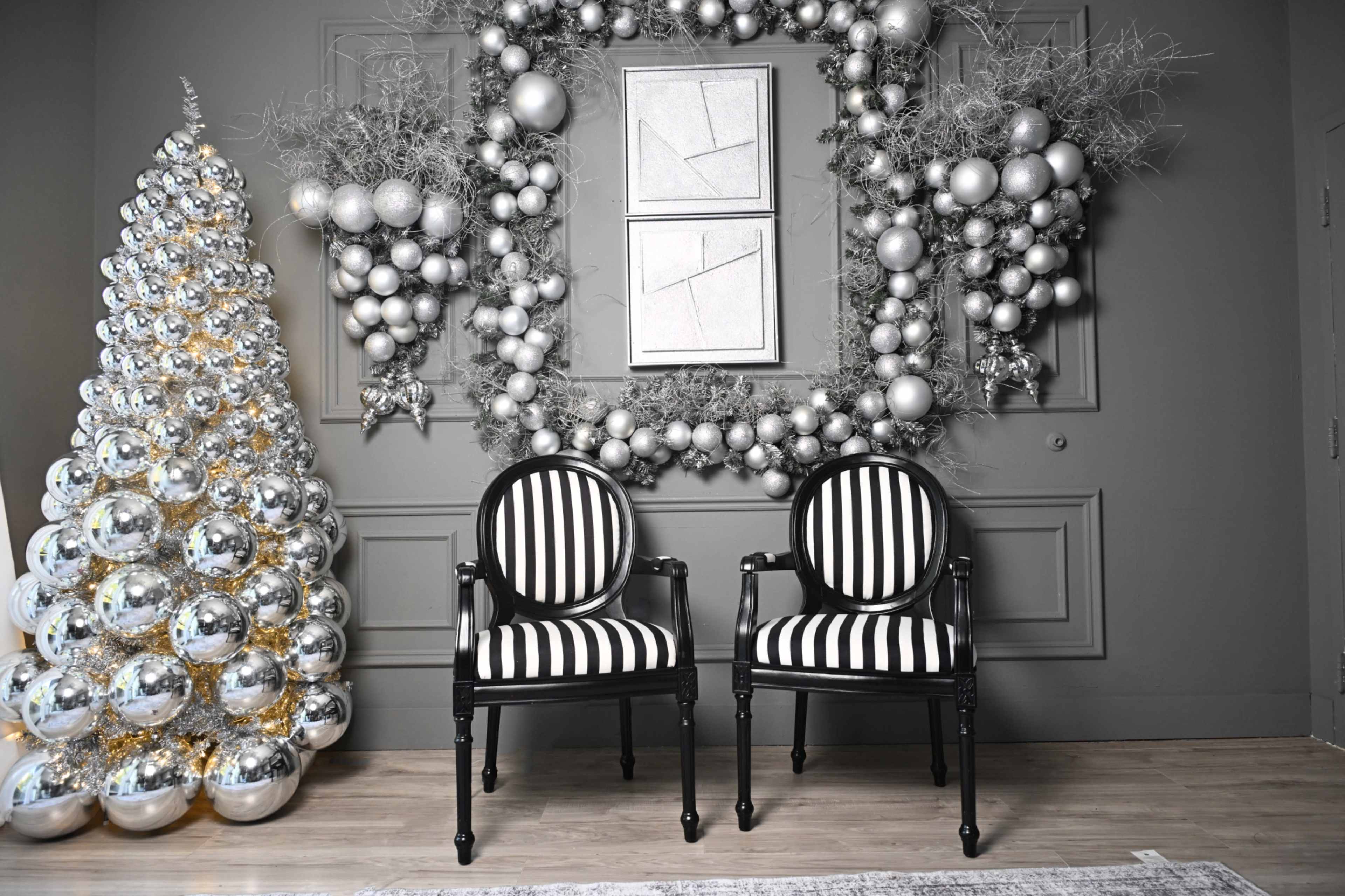The image depicts a festive indoor scene featuring two black and white striped chairs positioned in front of a wall adorned with a silver and white decorative arrangement, alongside a Christmas tree decorated with shiny ornaments.